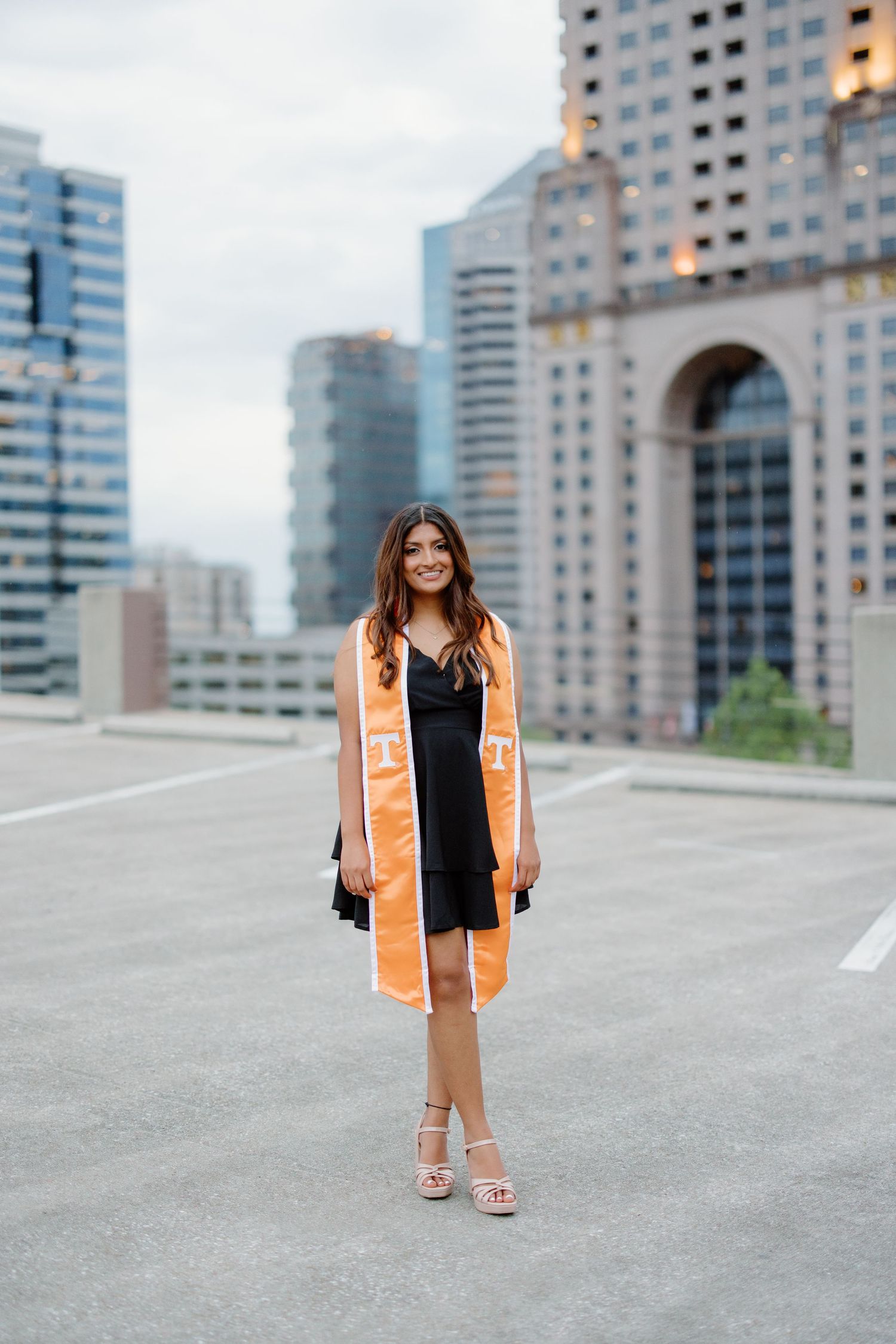 Someone poses in a black dress and orange coat against modern city buildings on a rooftop parking area.