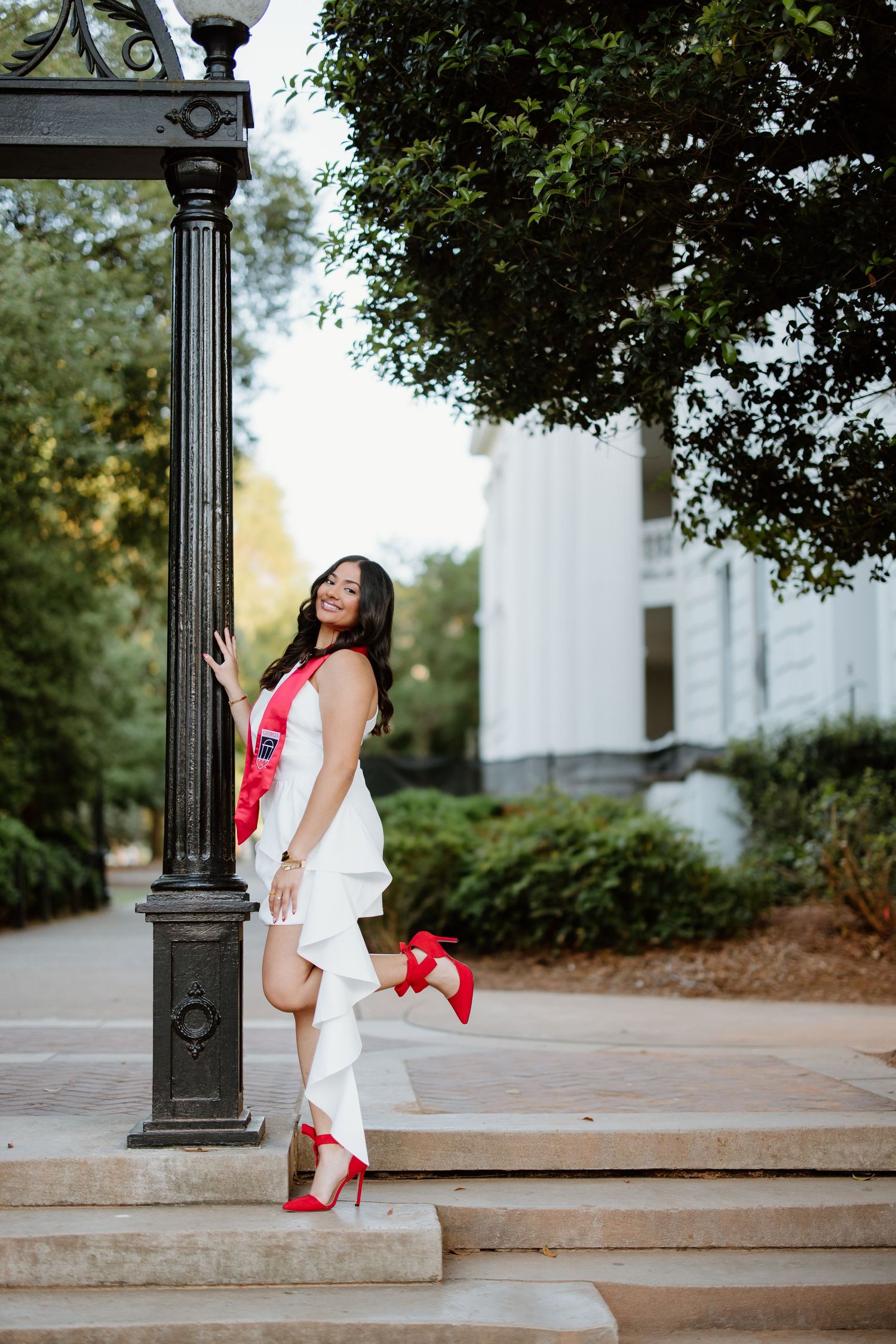 A person in a white dress and red heels poses playfully by a decorative lamp post on stone steps.