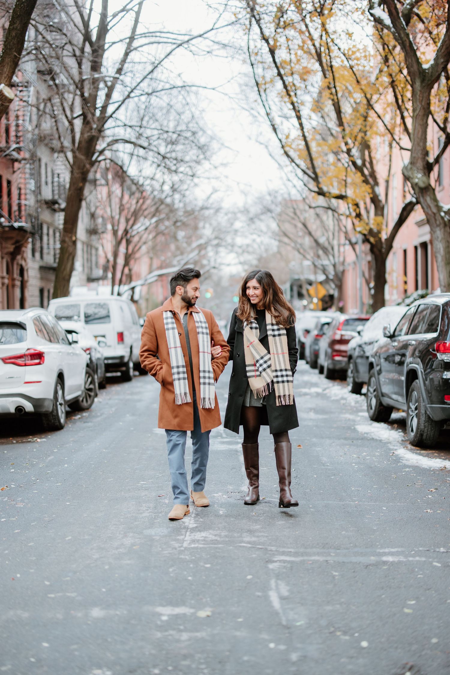 Two people walk together down a snowy residential street lined with parked cars and autumn trees.