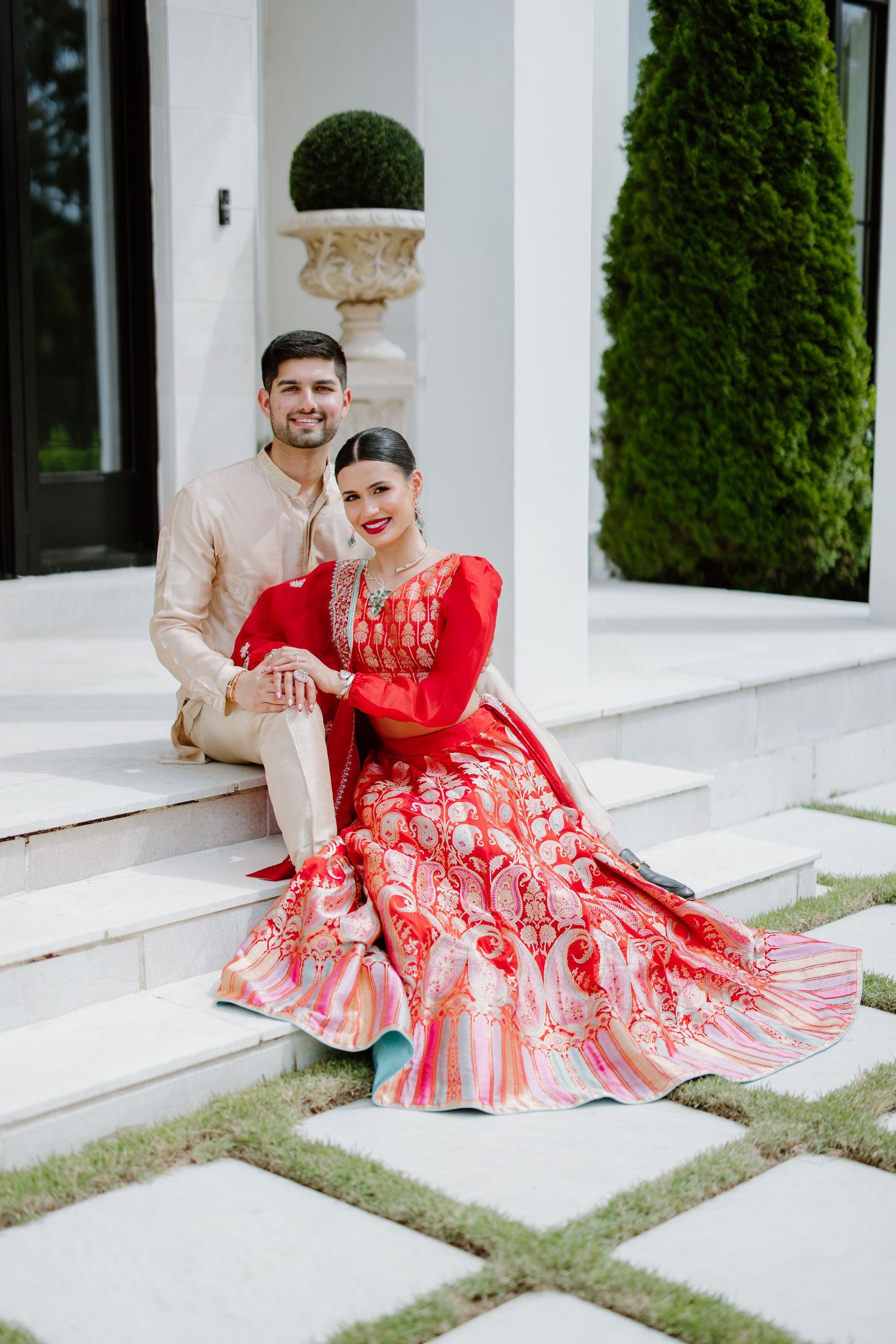 A couple in formal attire sits on white steps, with the woman wearing a stunning red lehenga with embroidered details.