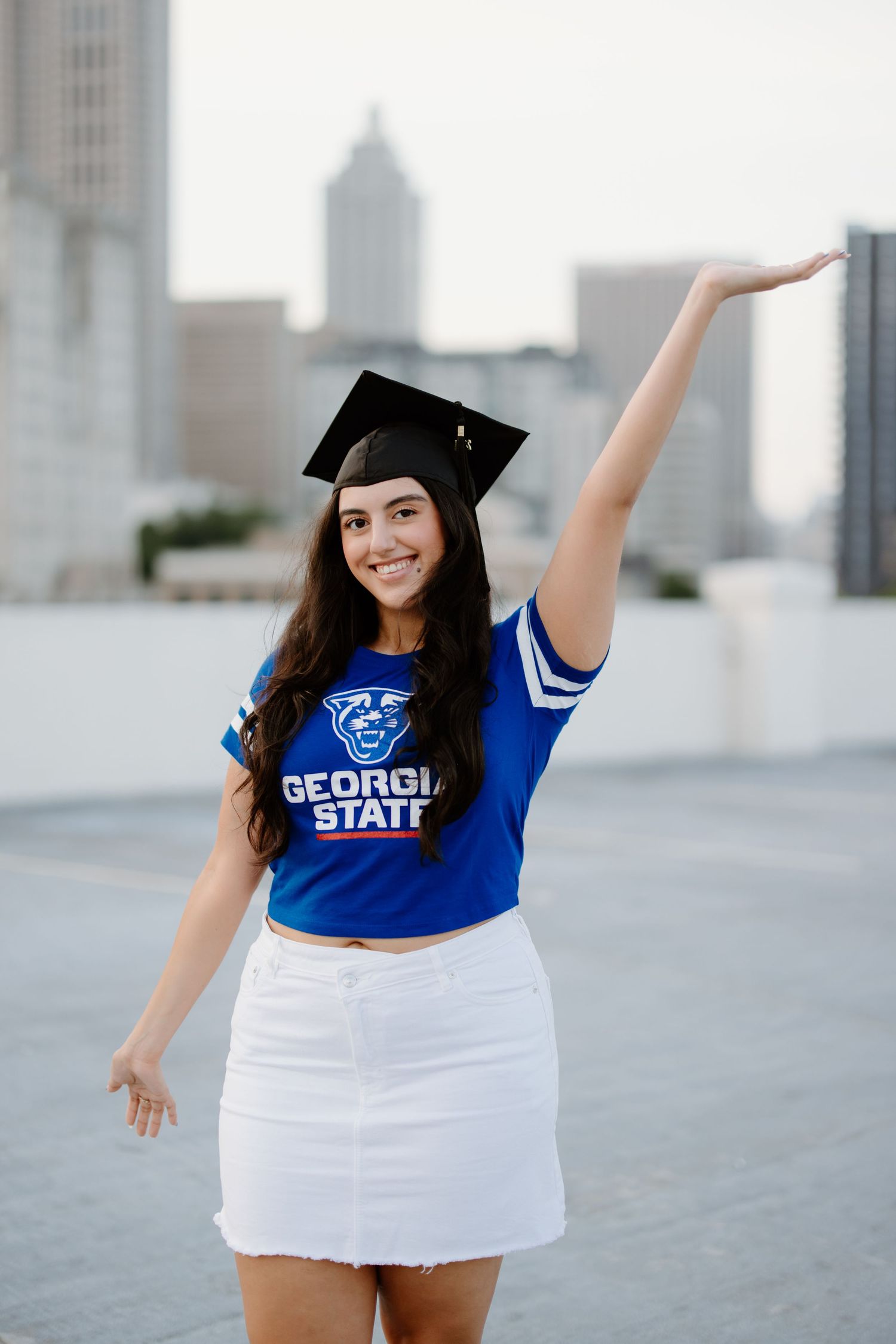Graduate in cap and gown wearing Georgia State University shirt poses on rooftop with Atlanta skyline in background.