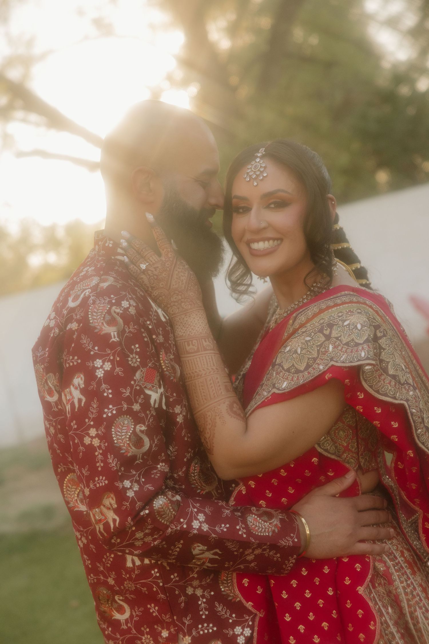 Couple in traditional red Indian wedding attire embrace outdoors, bride smiling in lehenga with gold jewelry and maang tikka.