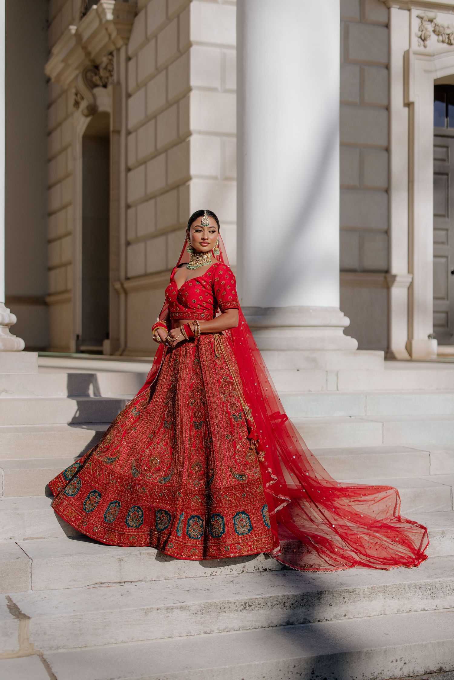 A traditional South Asian bride poses in an ornate red lehenga with trailing train against white columns.