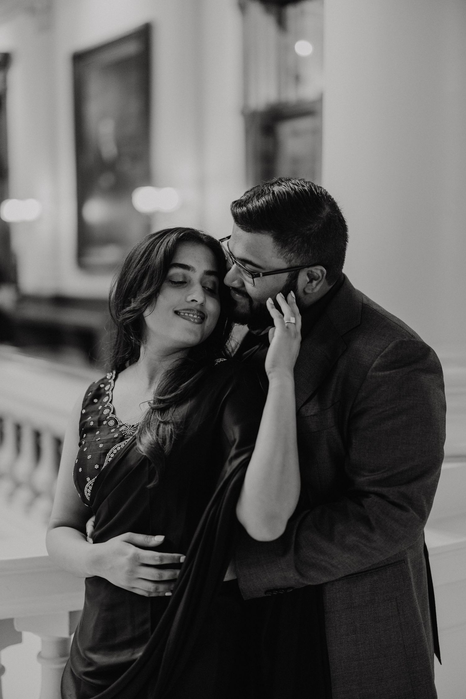 Black and white romantic portrait of a couple in formal attire embracing intimately against an elegant interior background.