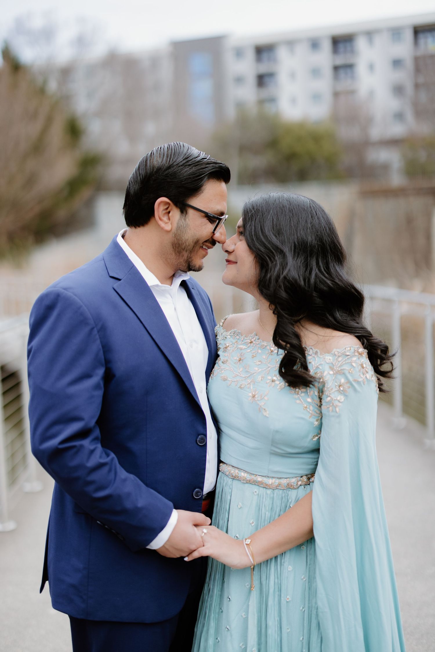 A couple in formal attire, with a navy suit and light blue flowing dress, share an intimate moment outdoors.