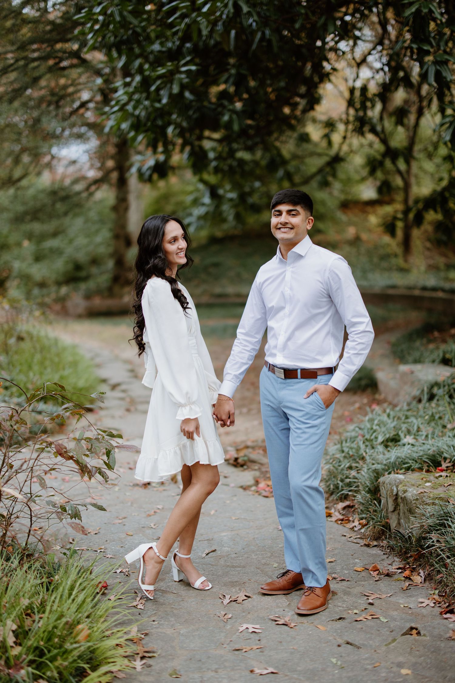 A couple in coordinated spring outfits hold hands while walking on a garden path surrounded by lush greenery.