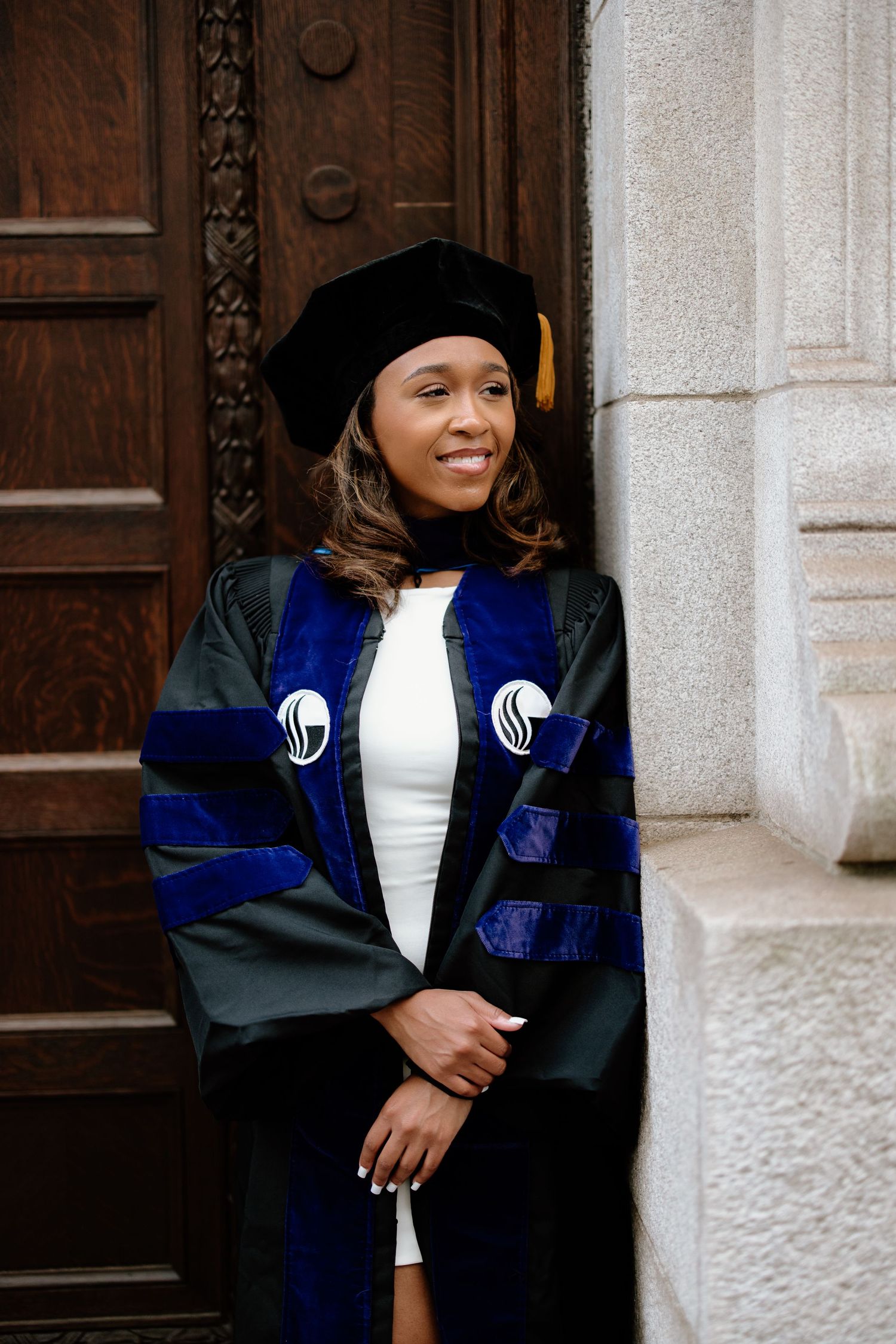 Graduate wearing a blue and black academic gown poses in front of a stone building entrance.