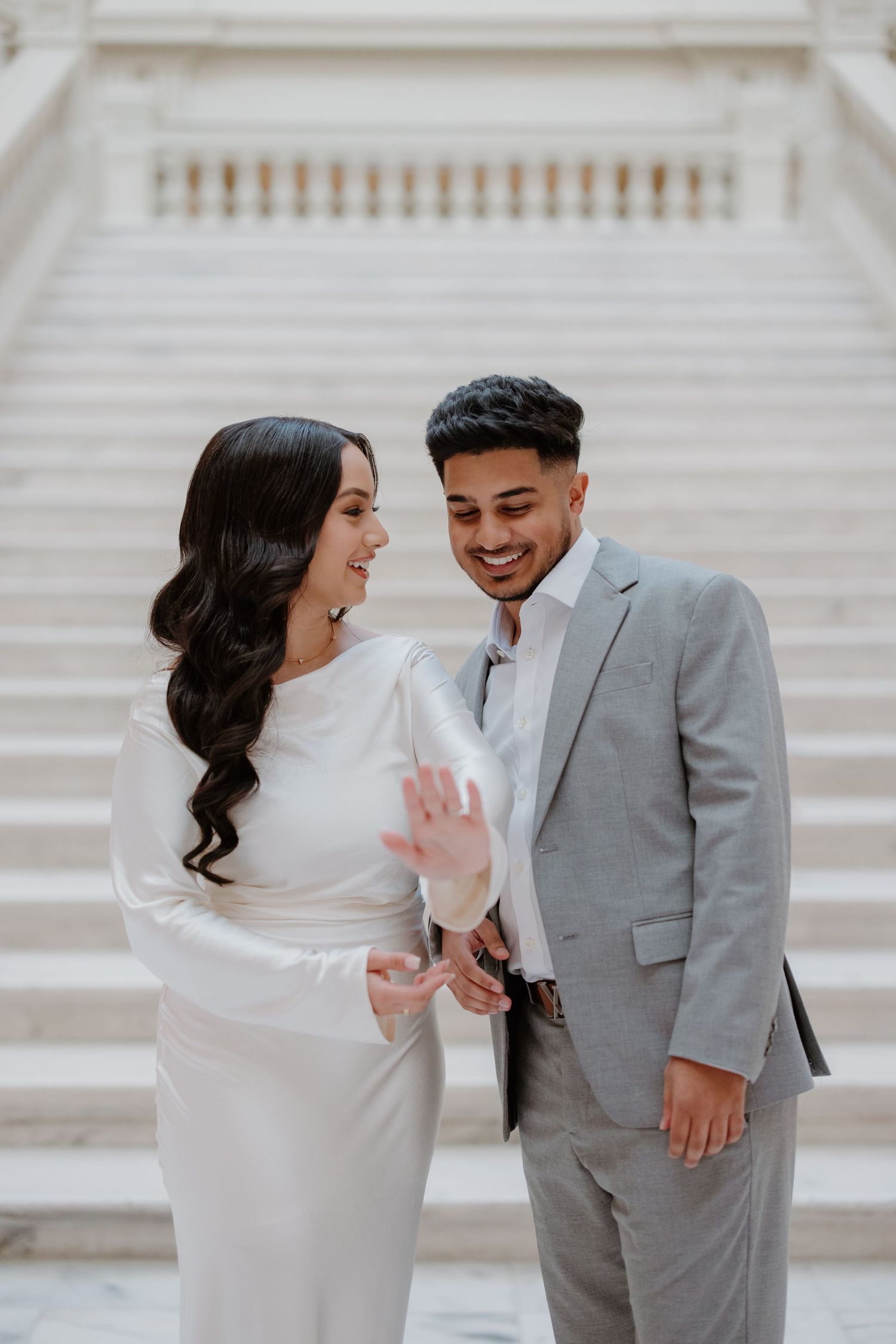 A couple shares a joyful moment together on white marble steps, wearing elegant gray and white formal attire.