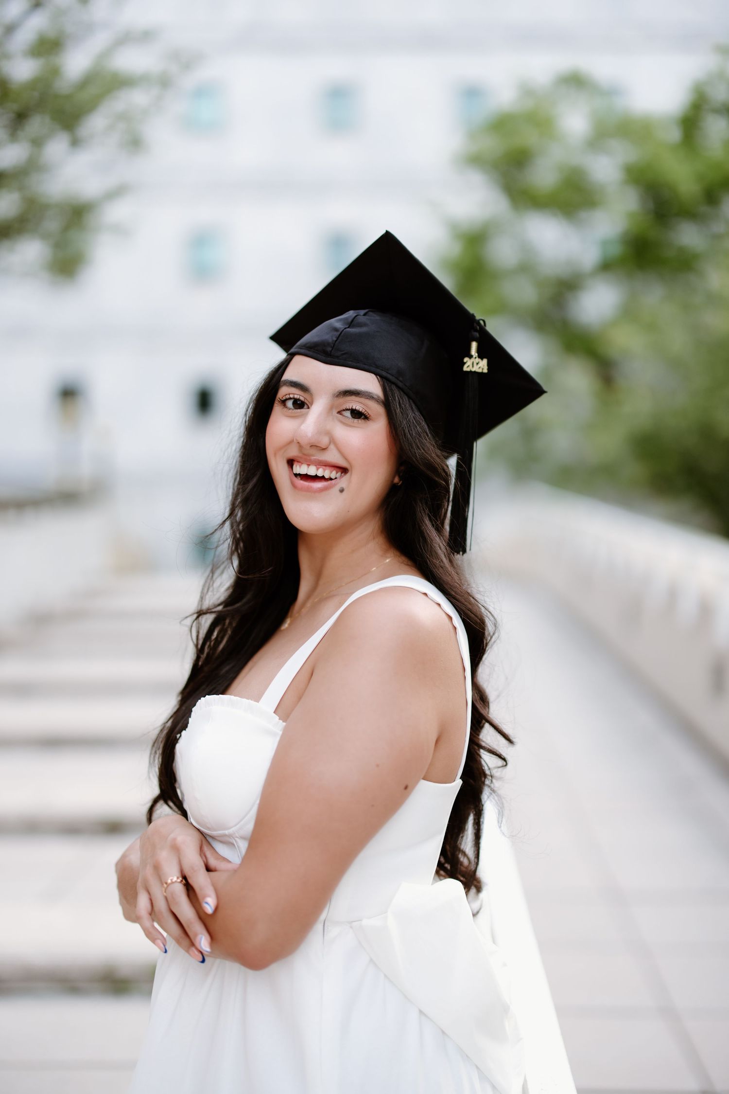Graduate in white dress and black mortarboard cap smiles while posing on outdoor walkway during graduation photoshoot.