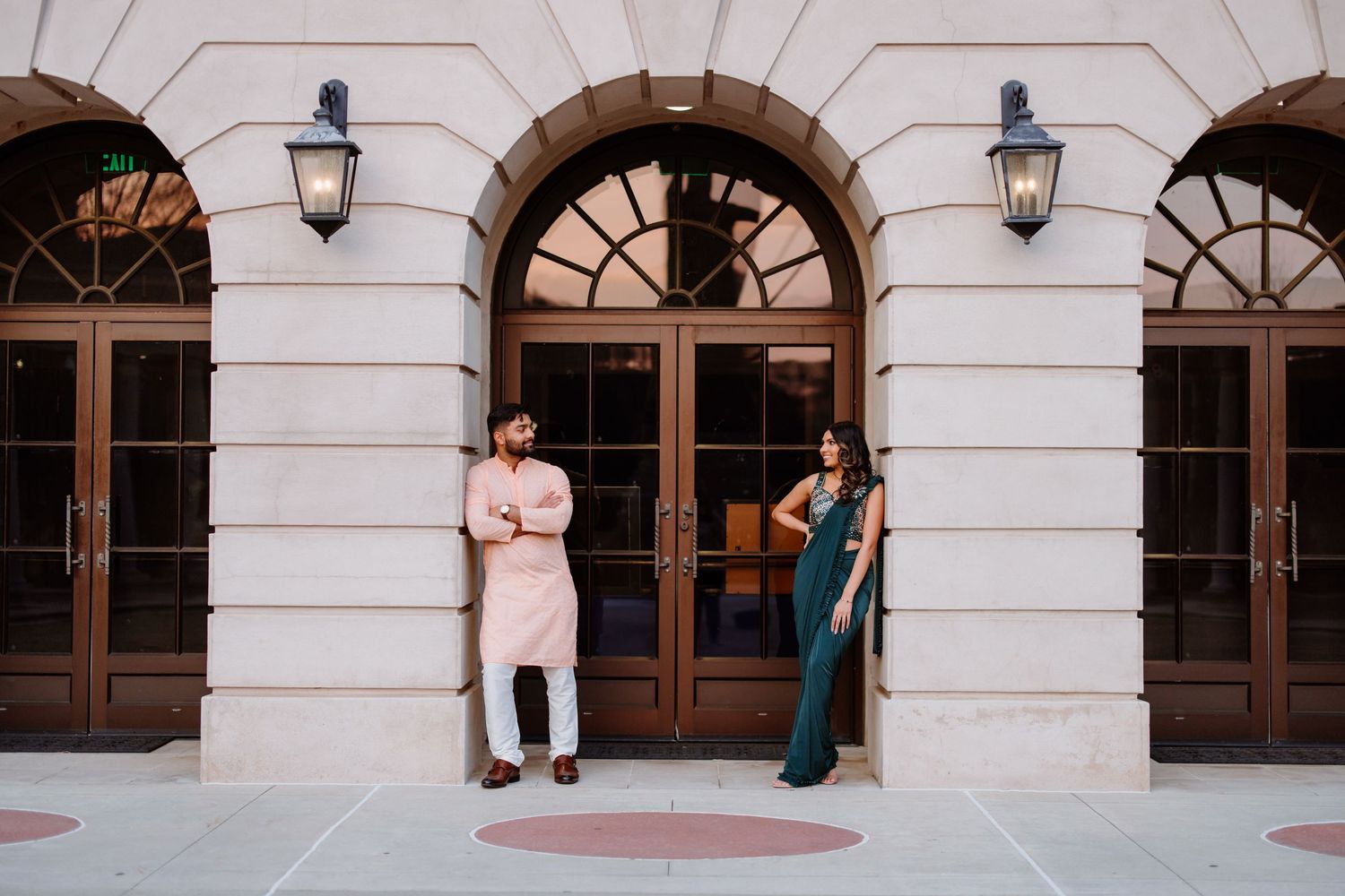 Two people stand in front of an elegant stone building with arched windows and wall sconces in an architectural setting.