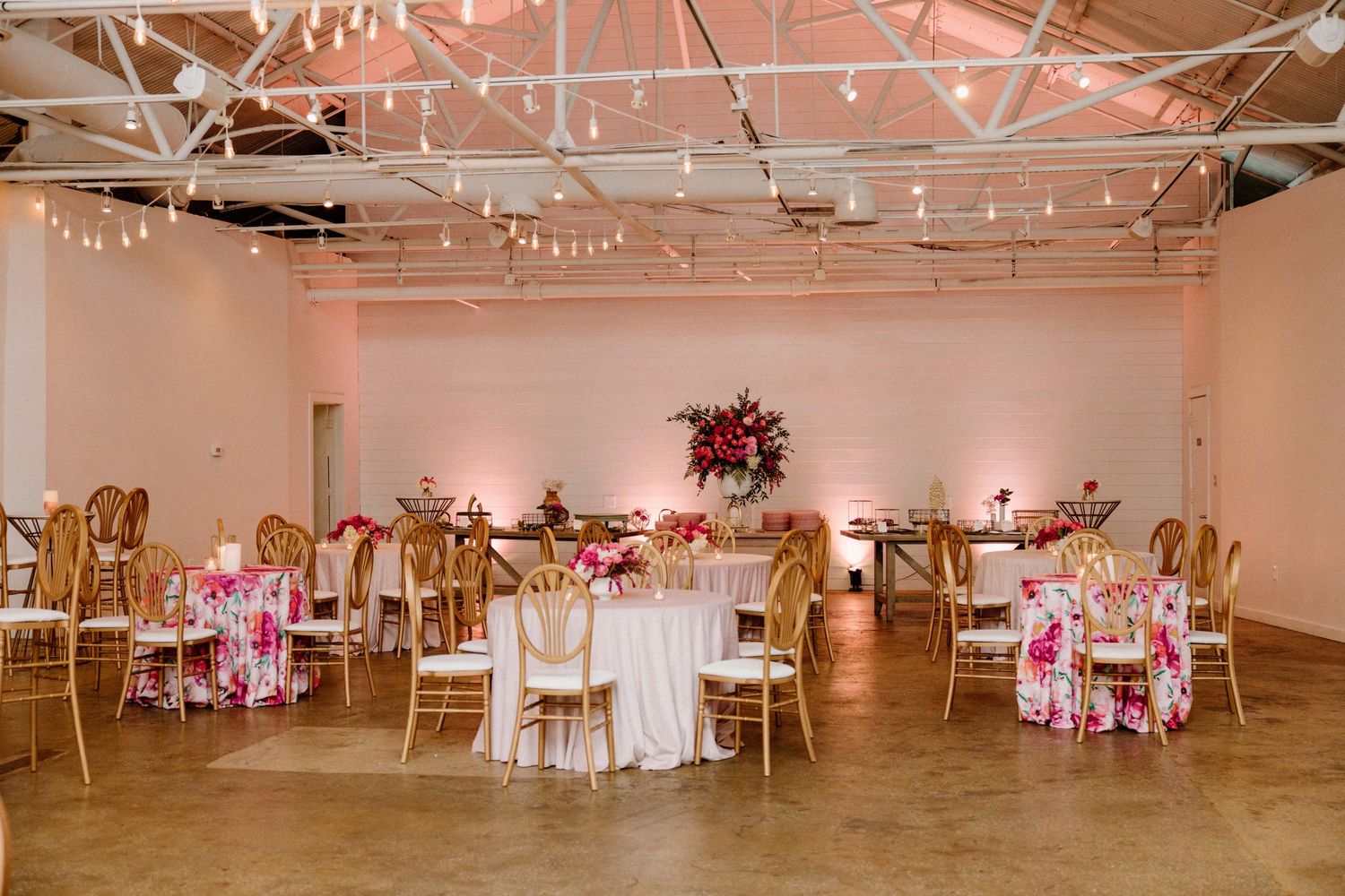 An elegant wedding reception venue with white and pink decor, gold chairs, and string lights hanging from the ceiling.