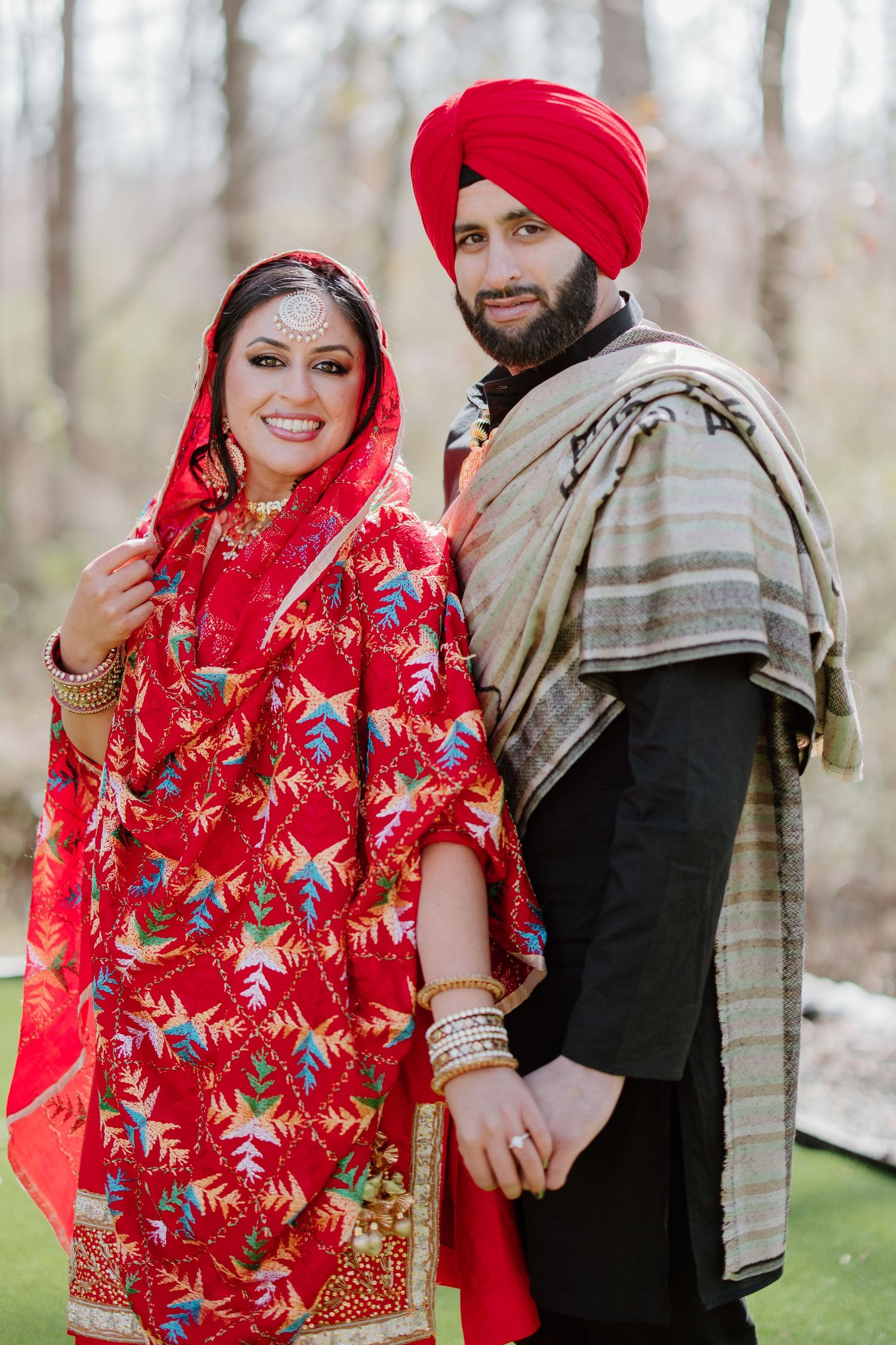A couple in traditional Indian wedding attire poses in a winter outdoor setting wearing vibrant red and black clothing.