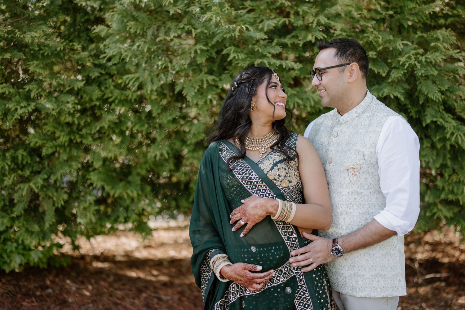 A couple stands close together in front of green foliage wearing formal traditional clothing at an outdoor photoshoot.