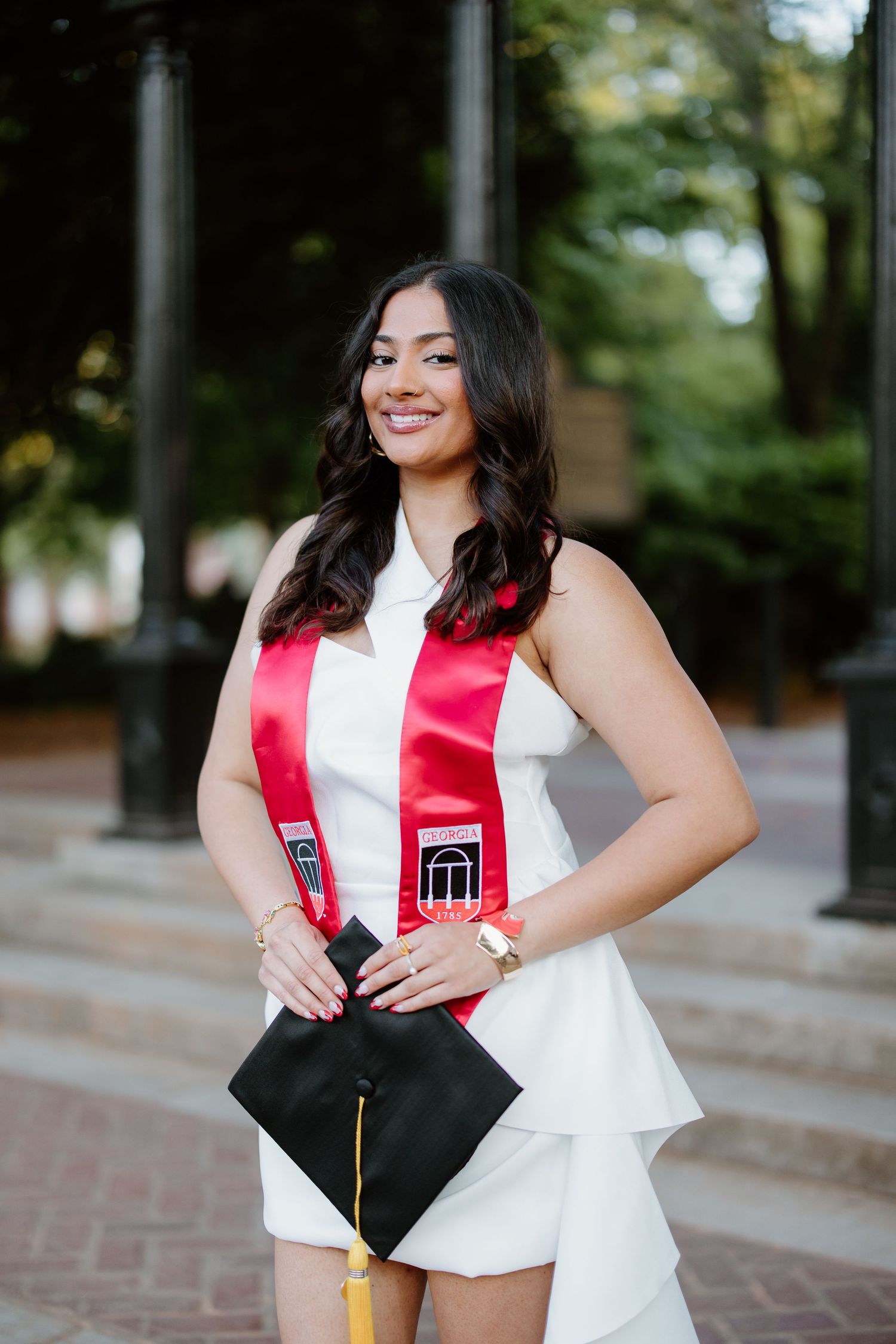 Graduate wearing white dress and red graduation stole holds black cap with tassel on brick walkway outdoors.