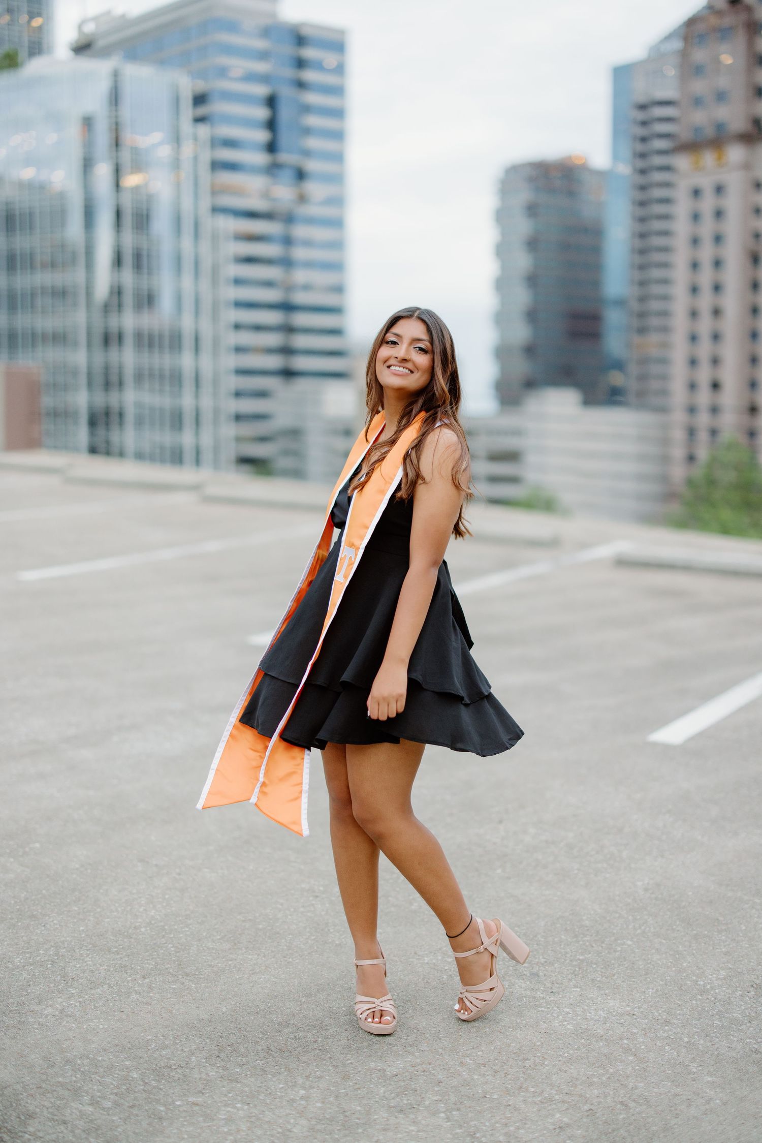 A fashionable outfit with a black dress, coral scarf and nude heels against an urban cityscape background.