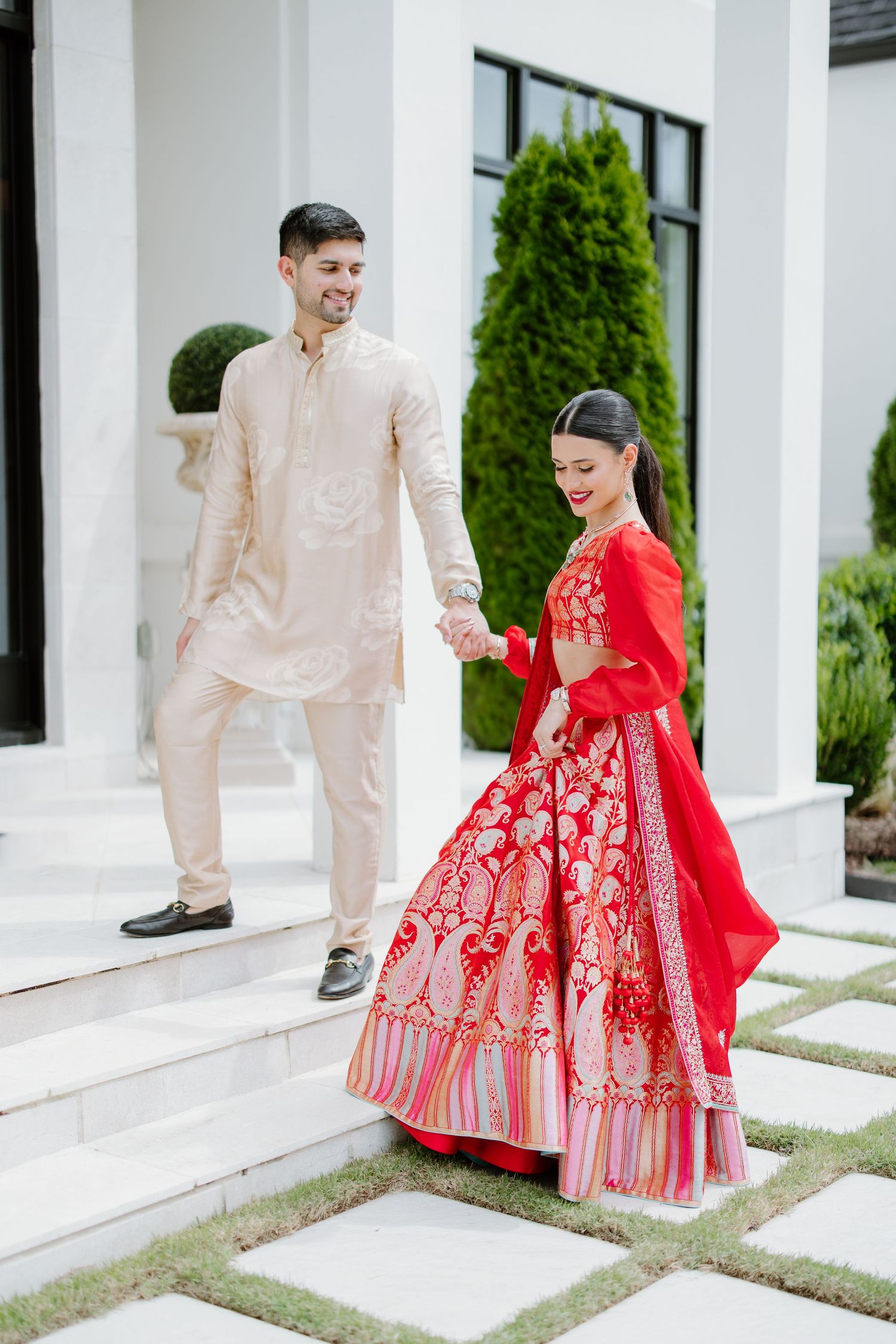 A couple in traditional fusion wedding attire poses on white steps, with the bride in a red embroidered gown.