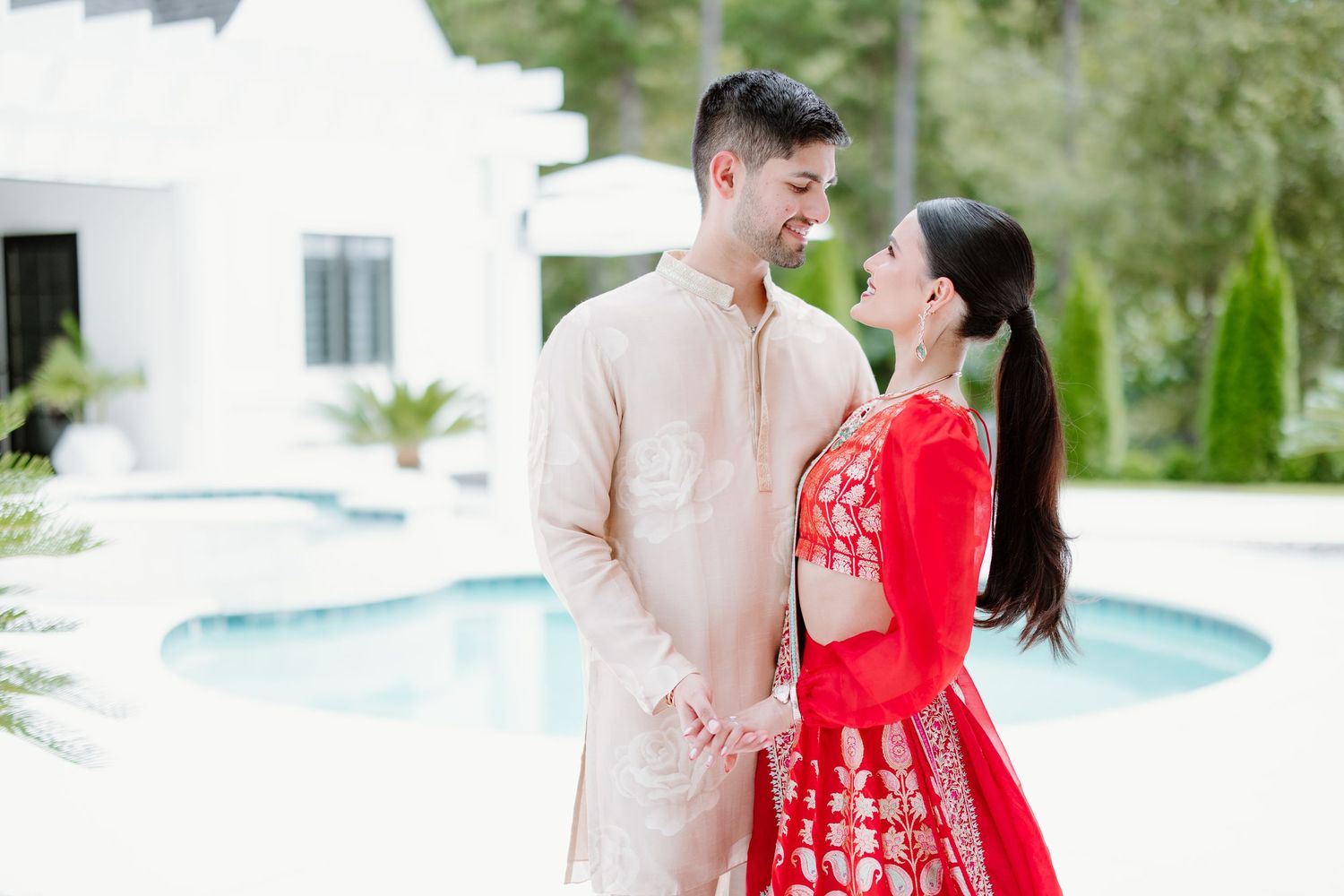 A couple poses together outdoors, with one wearing a red outfit and the other in beige, against a natural backdrop.