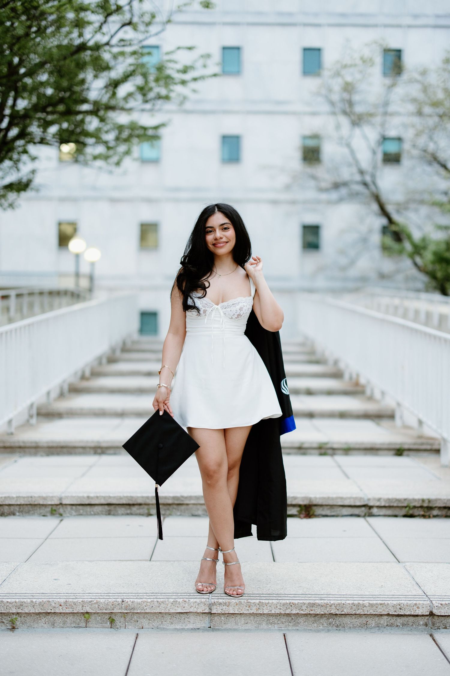 A person in a white sundress and heels poses on outdoor steps while holding graduation attire and accessories.