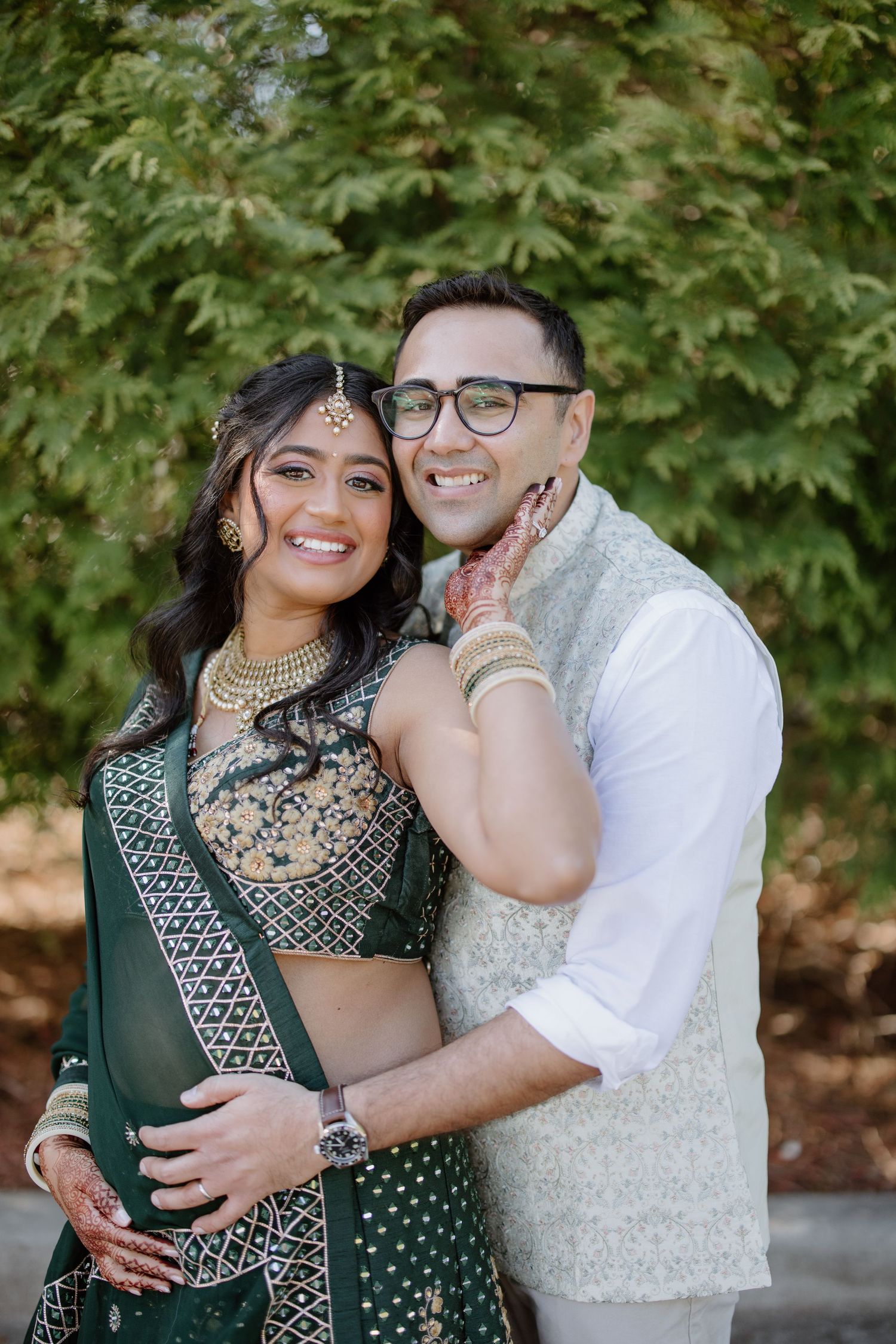 A couple in fusion wedding attire embrace outdoors, with the bride in an emerald green lehenga and traditional Indian jewelry.