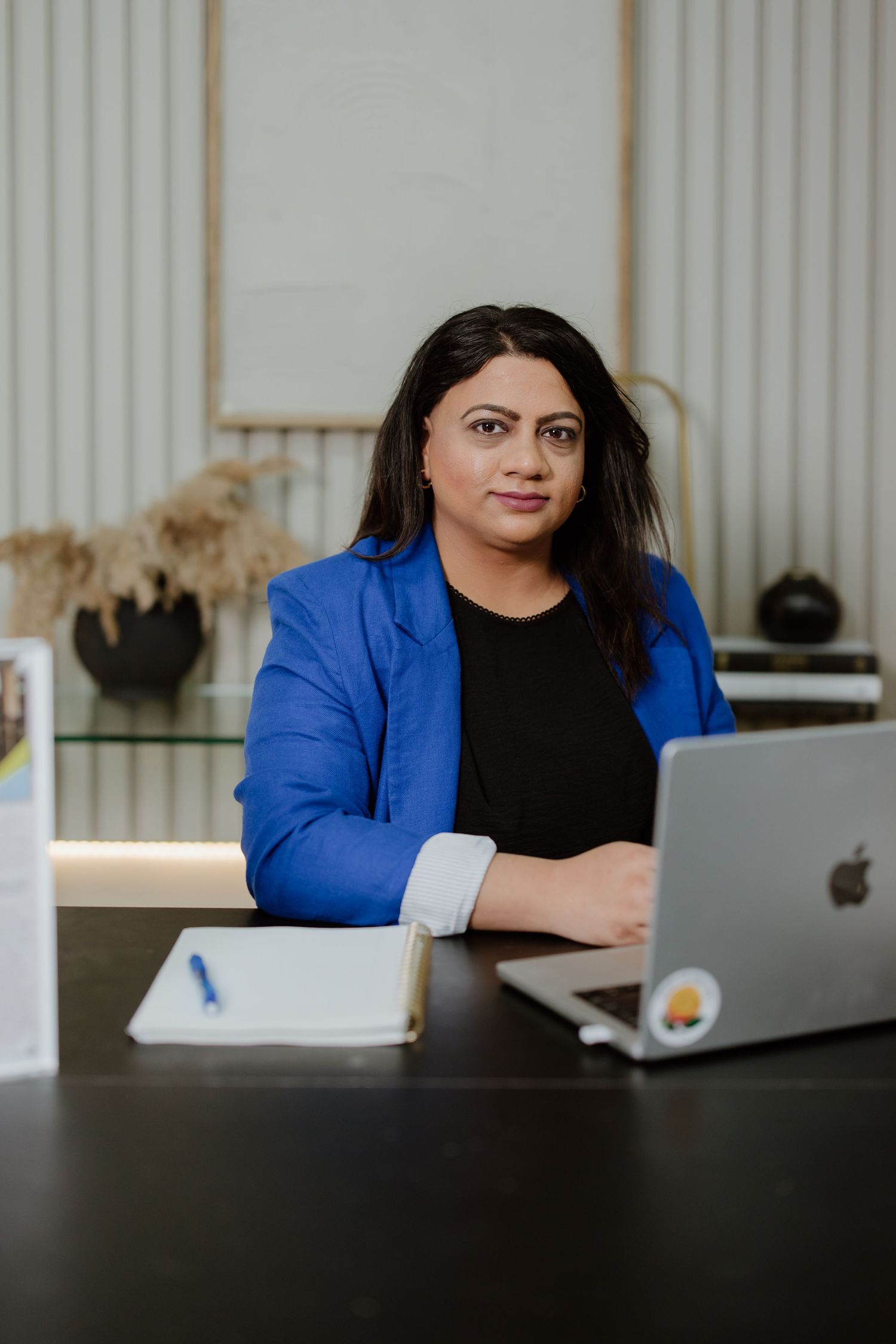 A woman in a bright blue blazer and black top sits at a desk with a MacBook laptop and notebook.