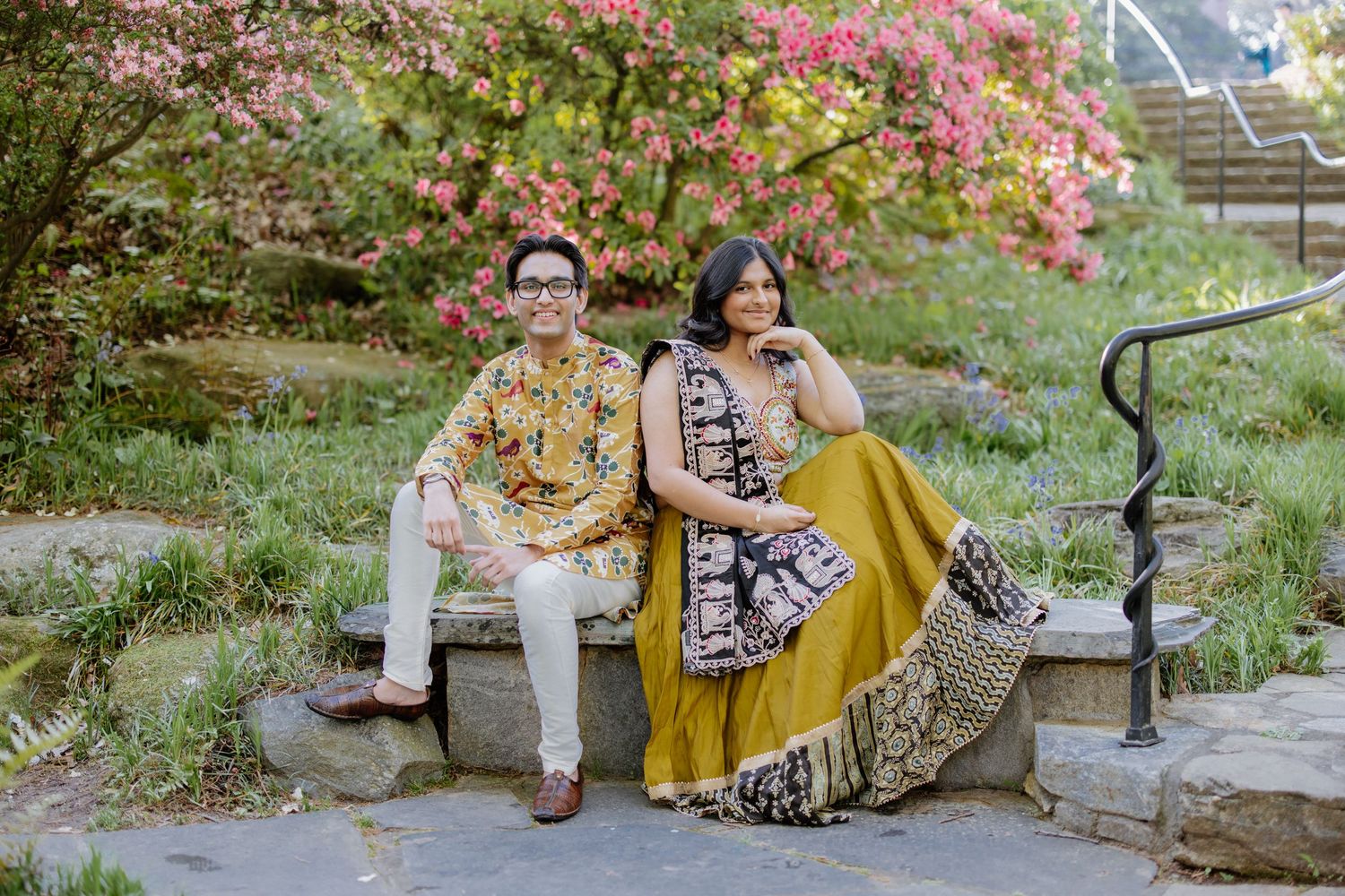 A couple in colorful traditional attire sits on stone steps surrounded by pink flowering bushes in a garden setting.