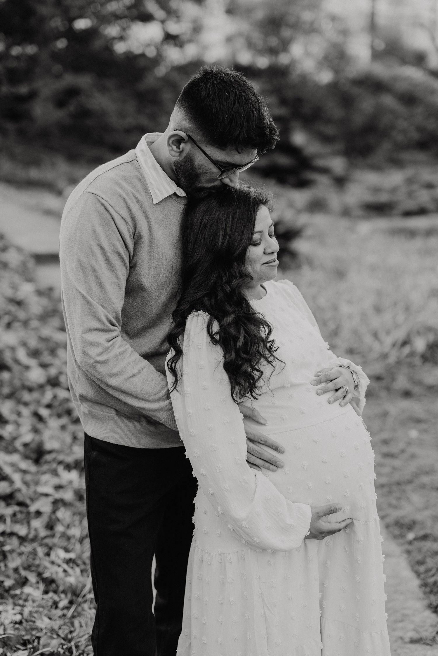 A tender black and white maternity photo shows a couple embracing outdoors in elegant white attire.