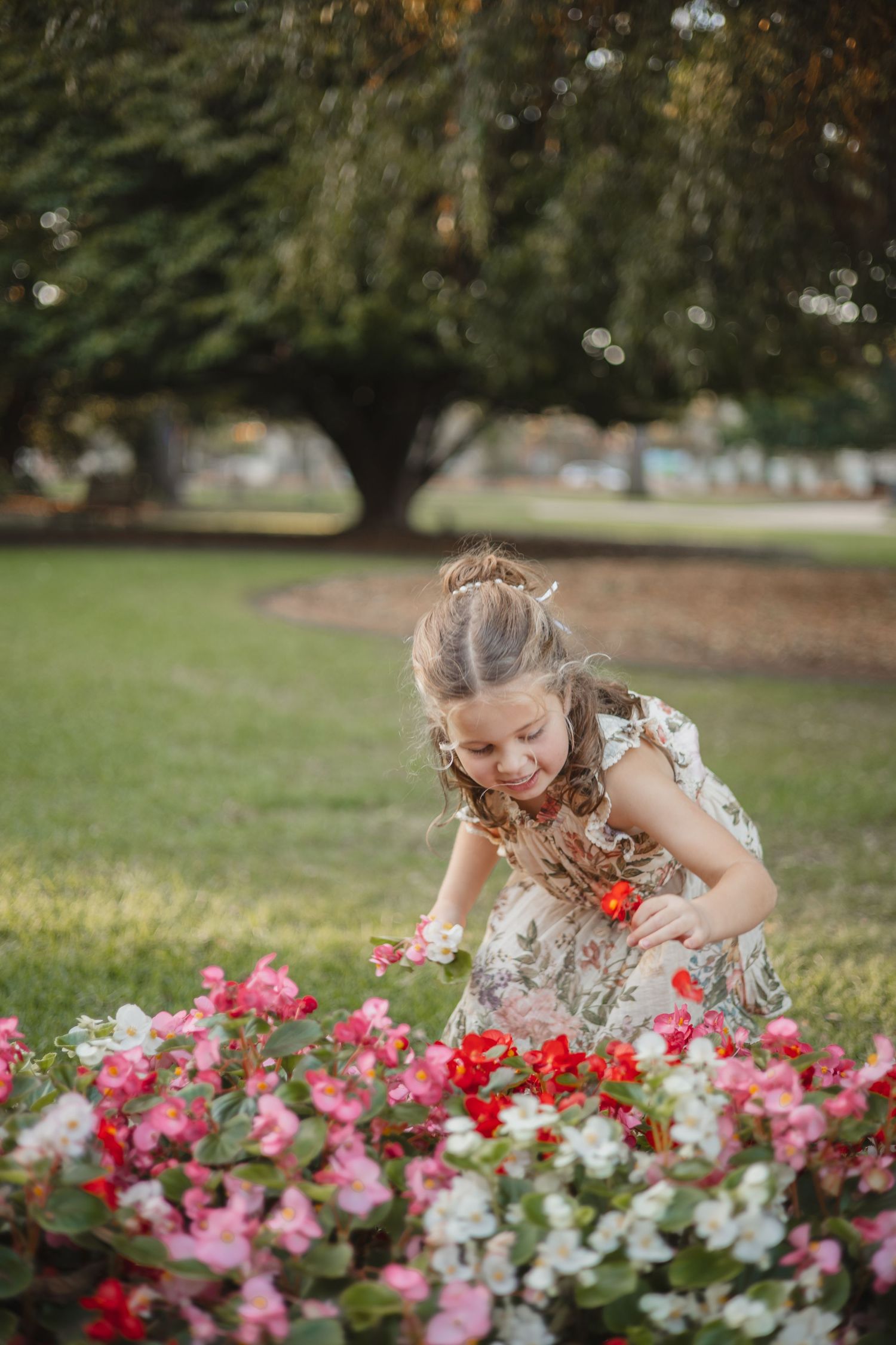 Young child in a floral dress bends down to admire pink and red flowers blooming in a garden on a sunny day.