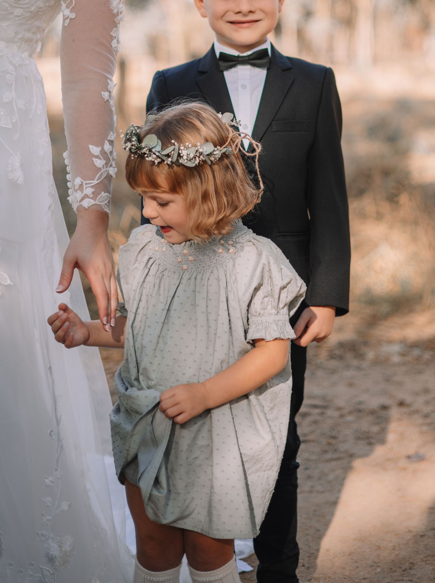 A small flower girl in a gray dress with a delicate floral crown stands between formal wedding attire at an outdoor ceremony.