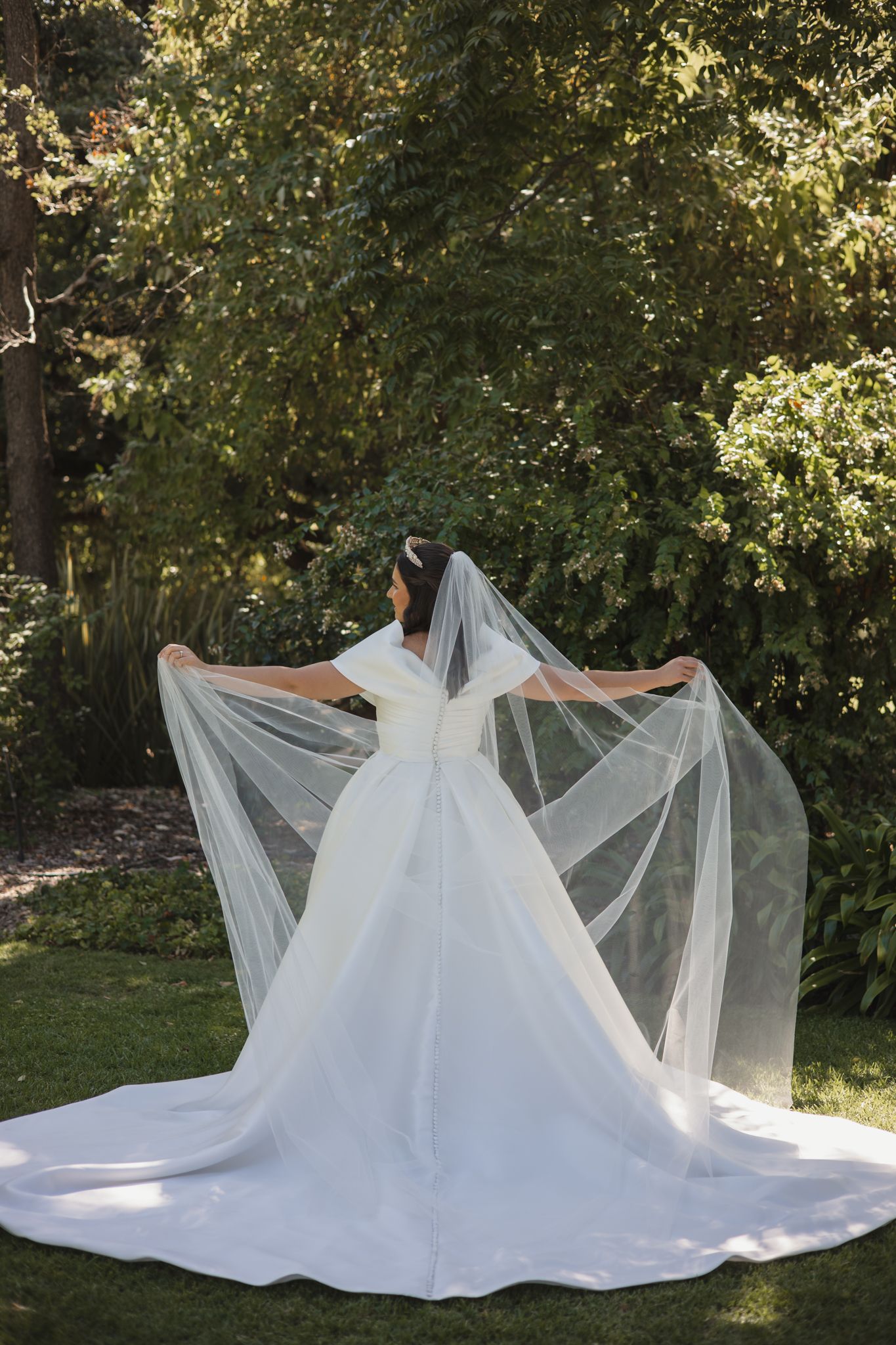 A flowing white wedding dress with a long veil laid out in a garden setting with sunlight filtering through trees.