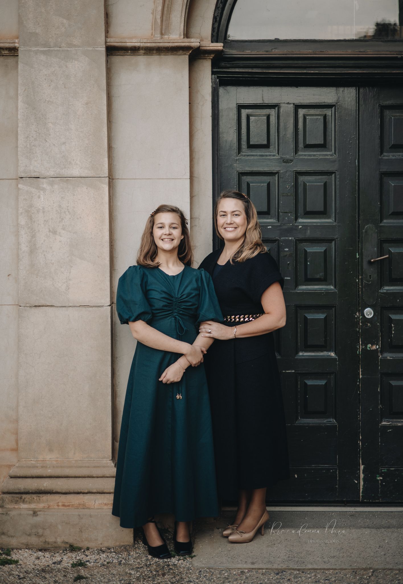 Mother and daughter wearing matching dark dresses pose together against a stone building with black doors.
