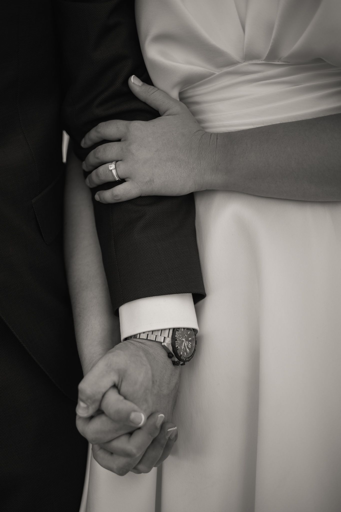 Black and white closeup of clasped hands showing wedding rings against formal attire.