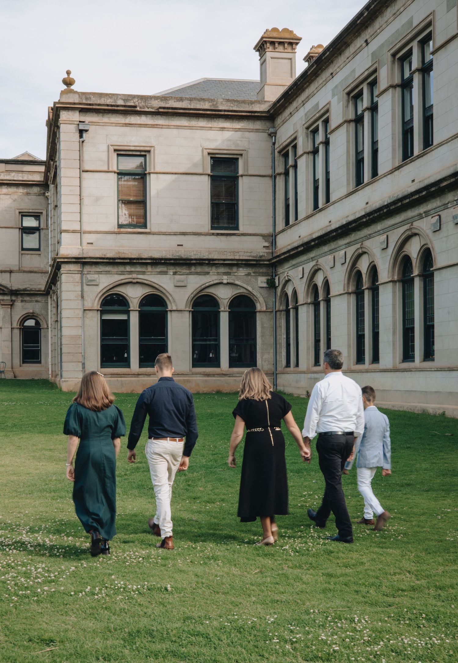 A group of people walking together on a grassy lawn in front of a grand historic stone building with arched windows.