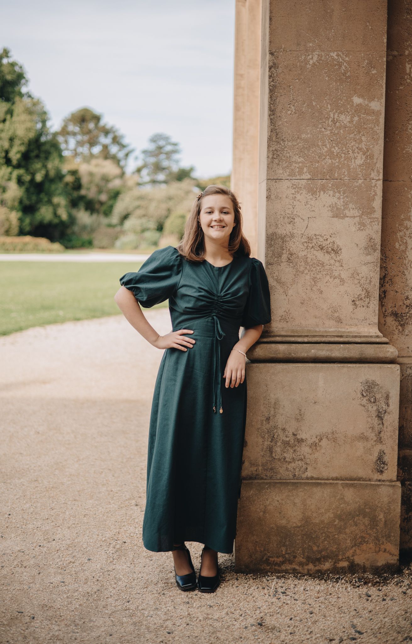 A person in a dark green dress poses against stone pillars in an outdoor setting.