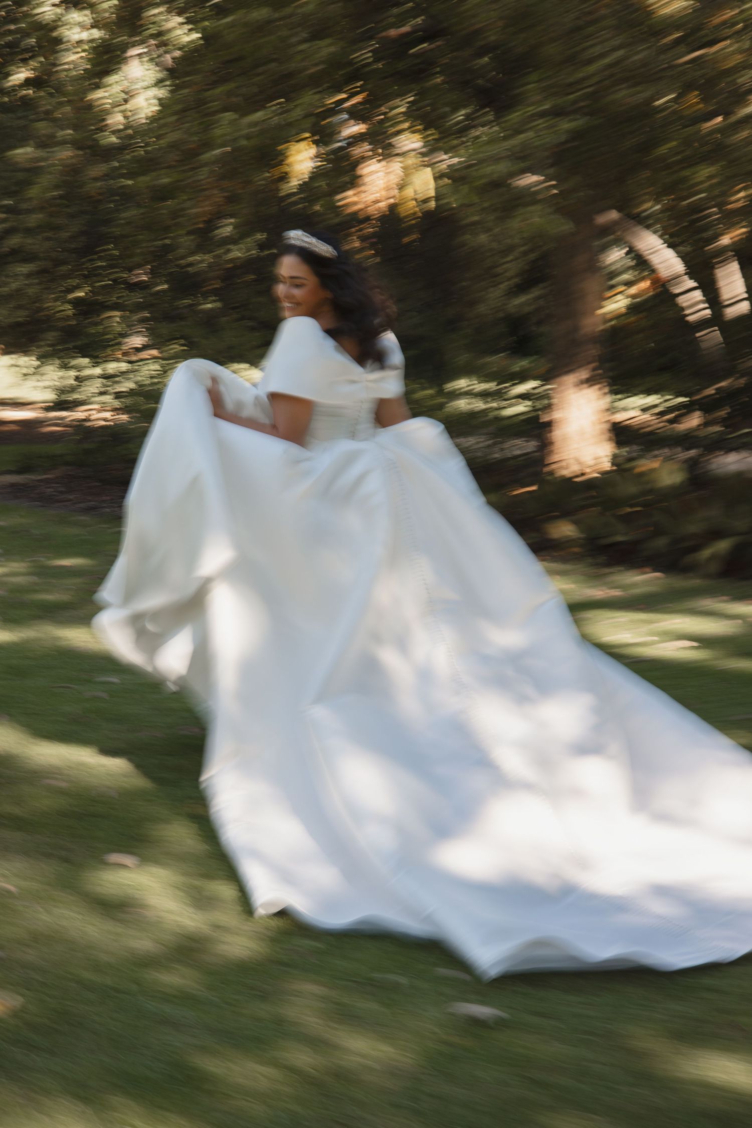 An elegant white wedding dress with off-shoulder design and flowing train photographed in motion on a sunny lawn.