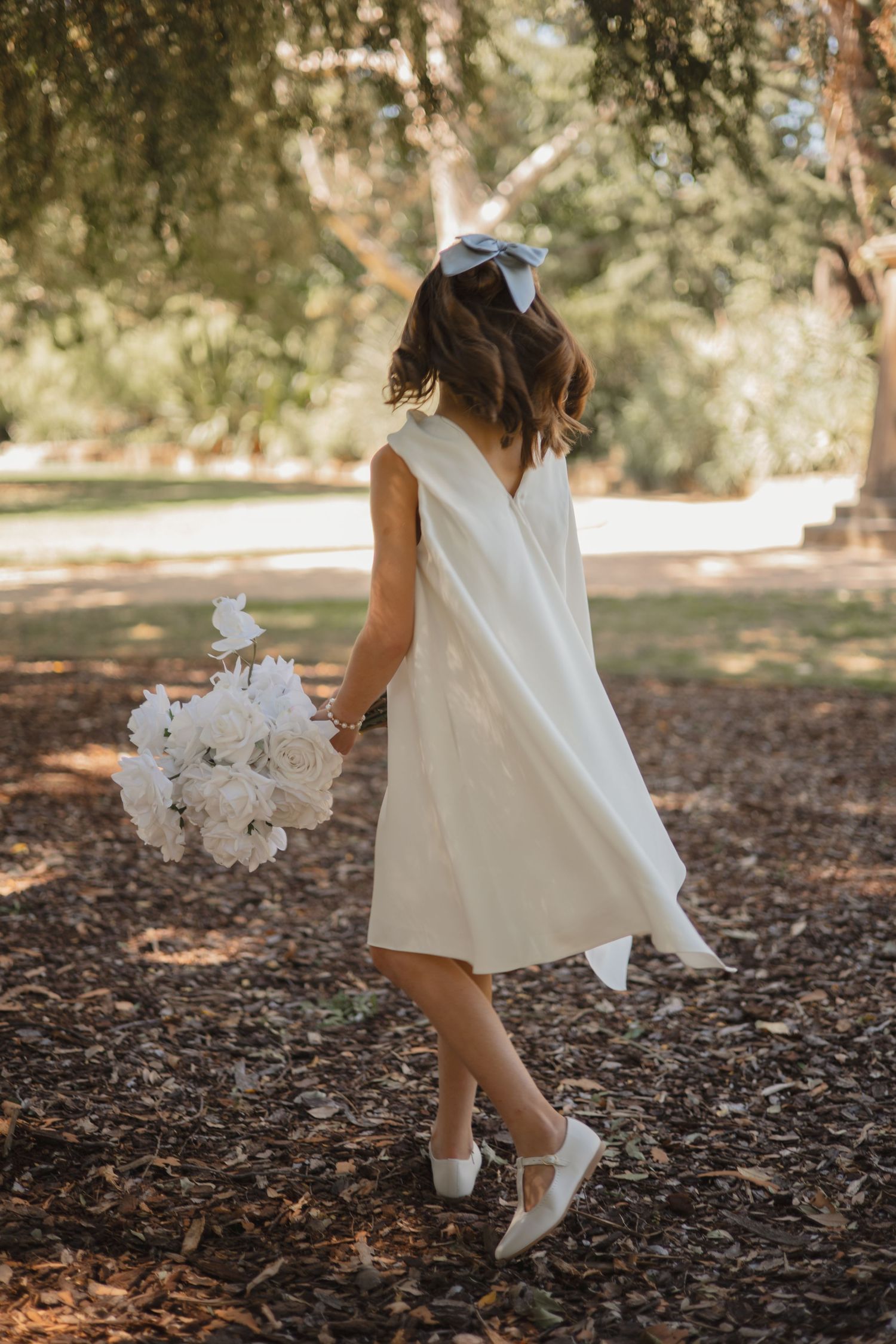 Person in white dress and bow holding flowers walks across woodchip-covered ground in a garden setting.