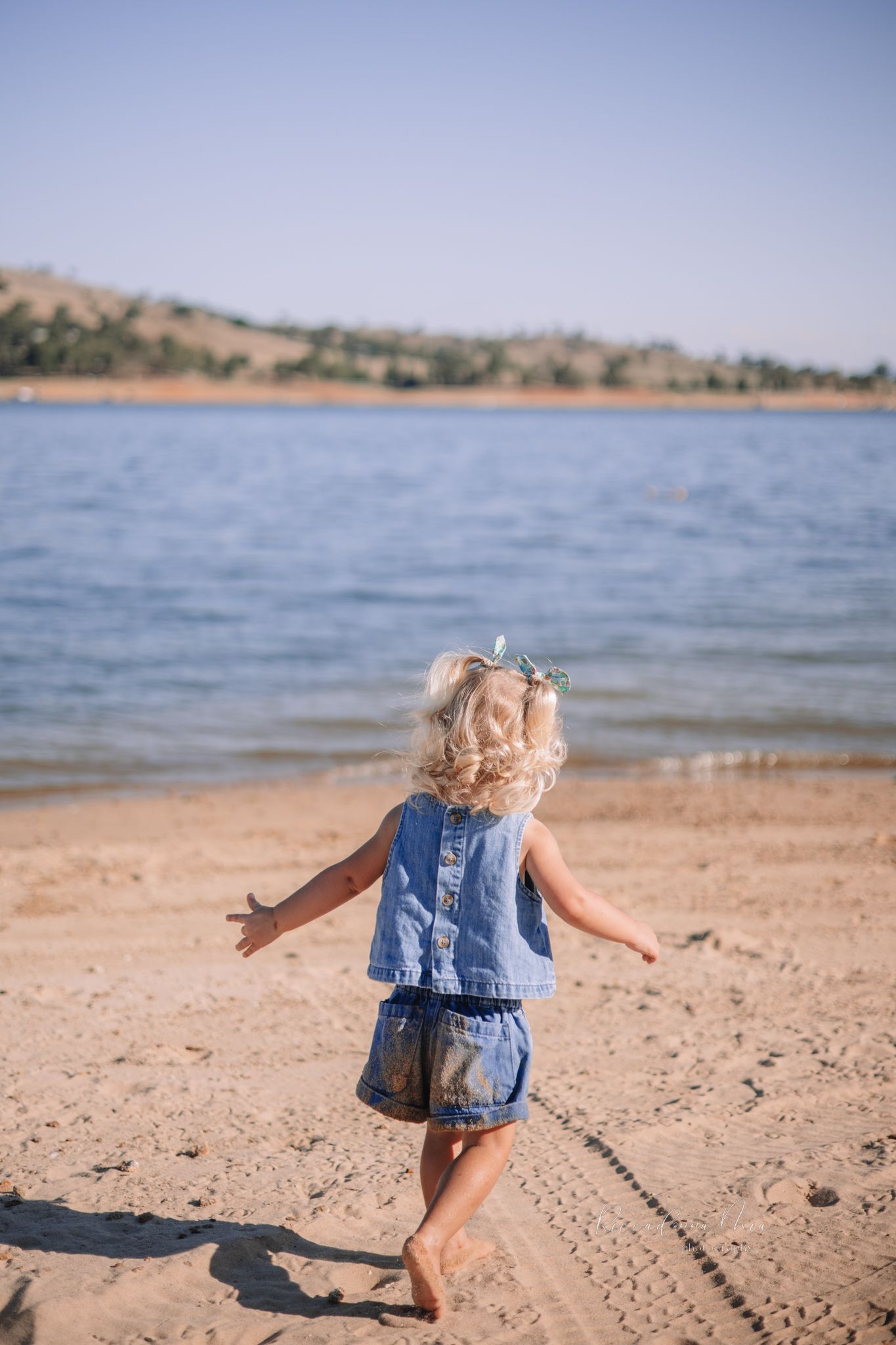 A small child runs playfully along a sandy lake shoreline on a sunny day.