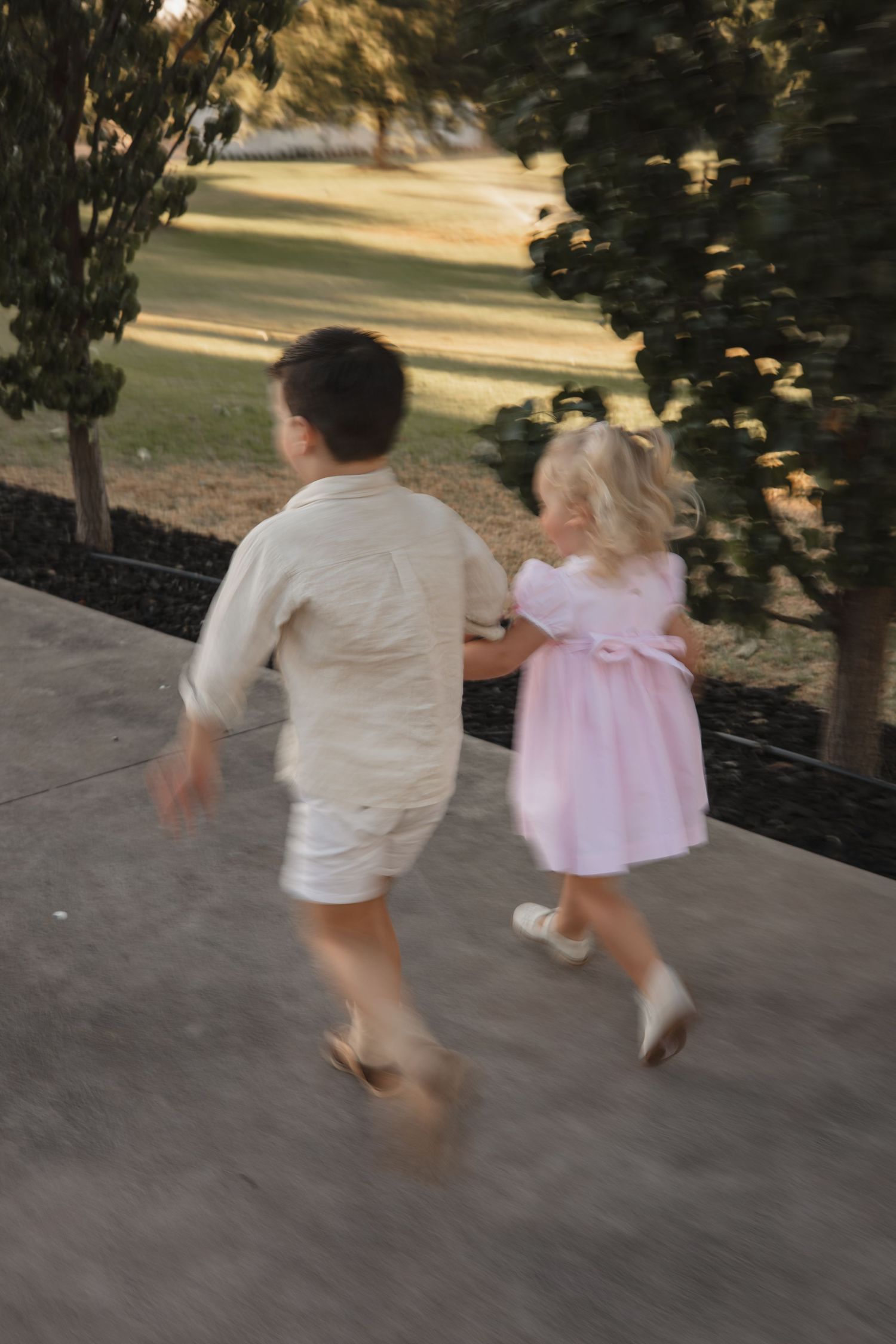 Two children running together on a concrete path, one wearing white and one in a pink dress, captured in motion at sunset.