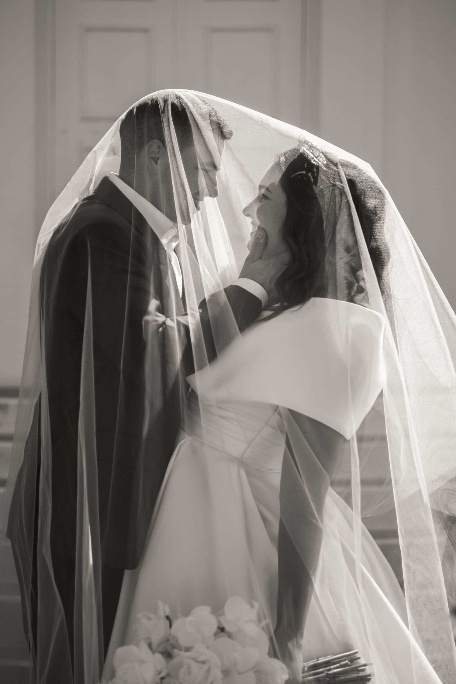 A romantic black and white wedding photo of a couple beneath a flowing veil during their ceremony.