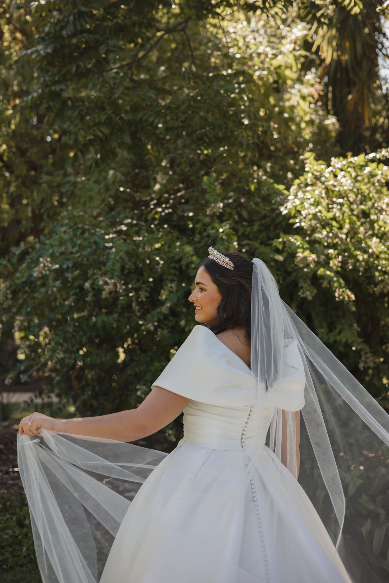 A bride in a classic white wedding dress with button details poses outdoors with a flowing veil in natural sunlight.
