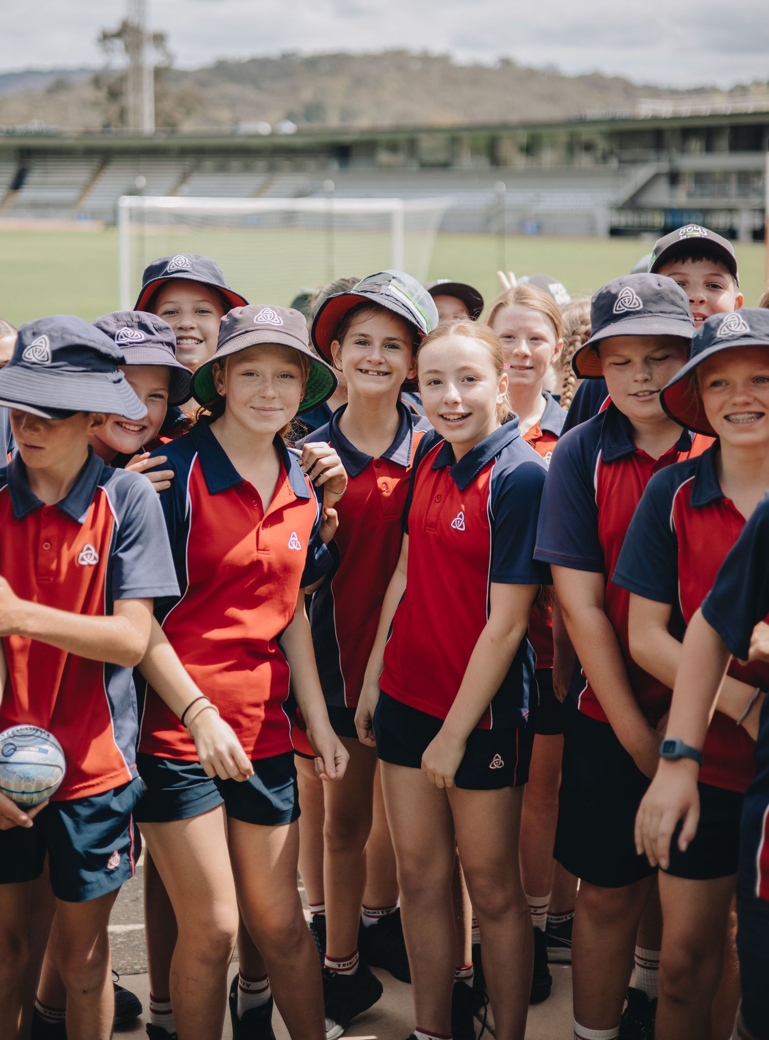 Students in red and navy school sports uniforms gather together on a sports field, wearing sun-protective hats.