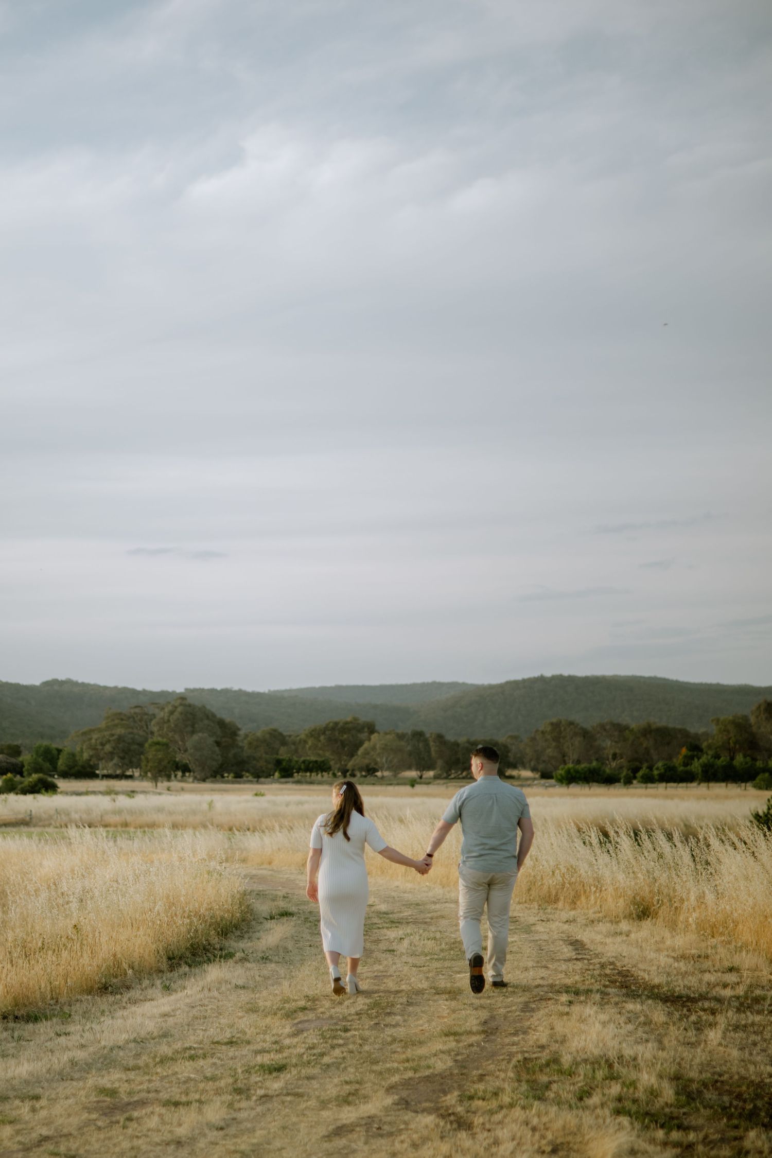 A family holds hands while walking together through a golden grass field against rolling hills on an overcast day.