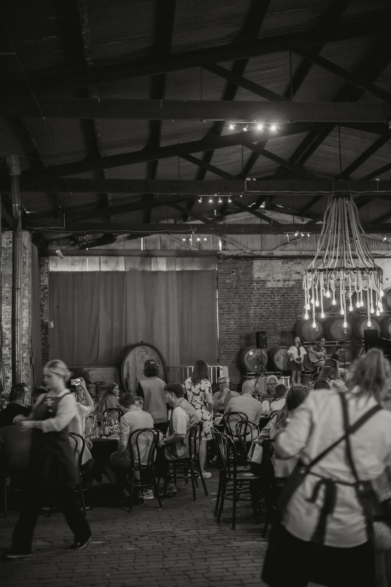 A black and white photo of a dimly lit restaurant interior with brick walls, chandeliers, and diners at tables at an event.