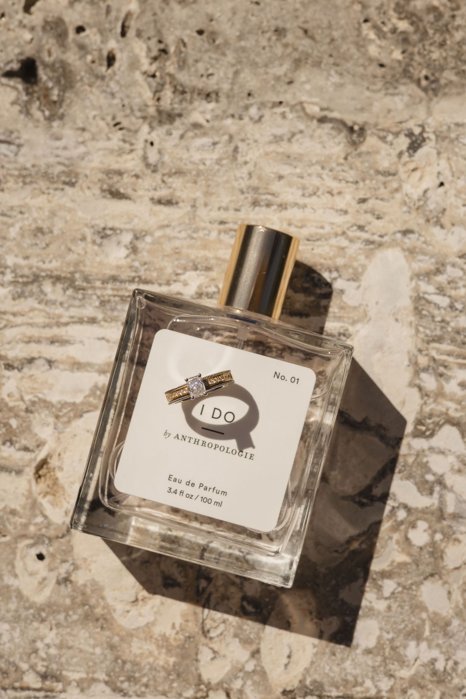 A square glass perfume bottle with gold cap labeled 'I Do' sits on a textured beige stone surface.