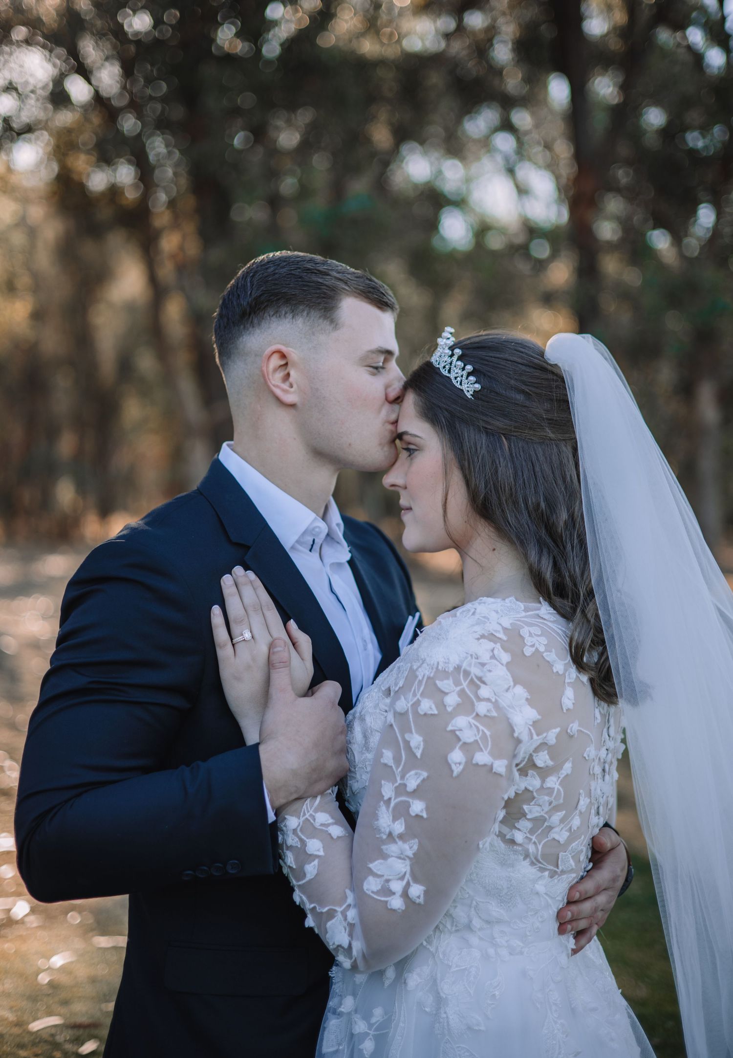 Tender romantic moment between newlyweds in woodland setting with bride in lace gown and veil, groom in navy suit.