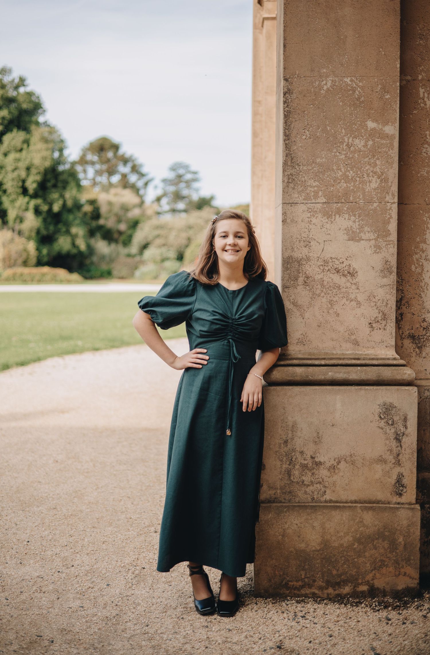 A woman in a vintage-style dark green dress poses next to a stone column in a garden setting.