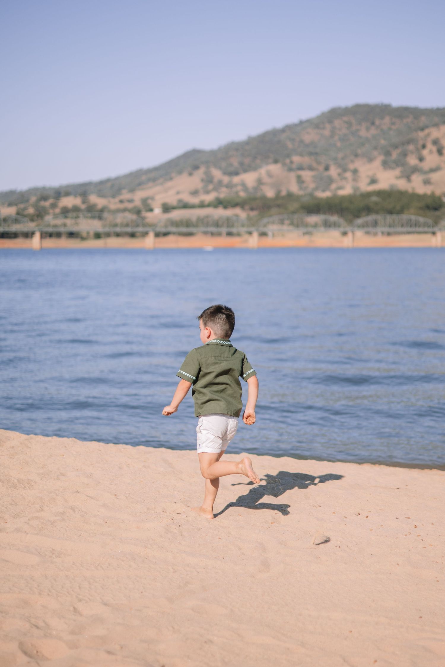 Small child in green shirt and white shorts runs along sandy beach shore with mountains and lake in background.
