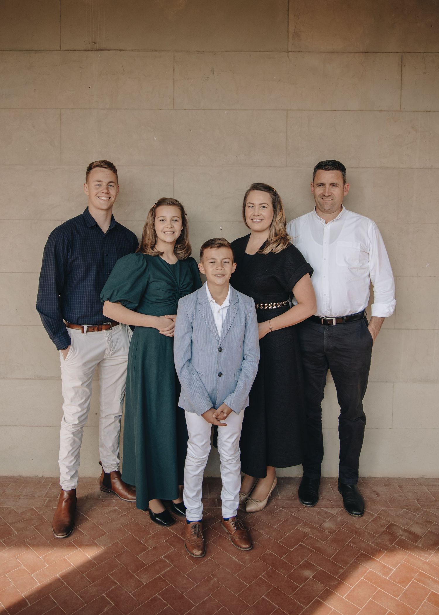 A group of five people in coordinated formal attire pose together against a beige stone wall.