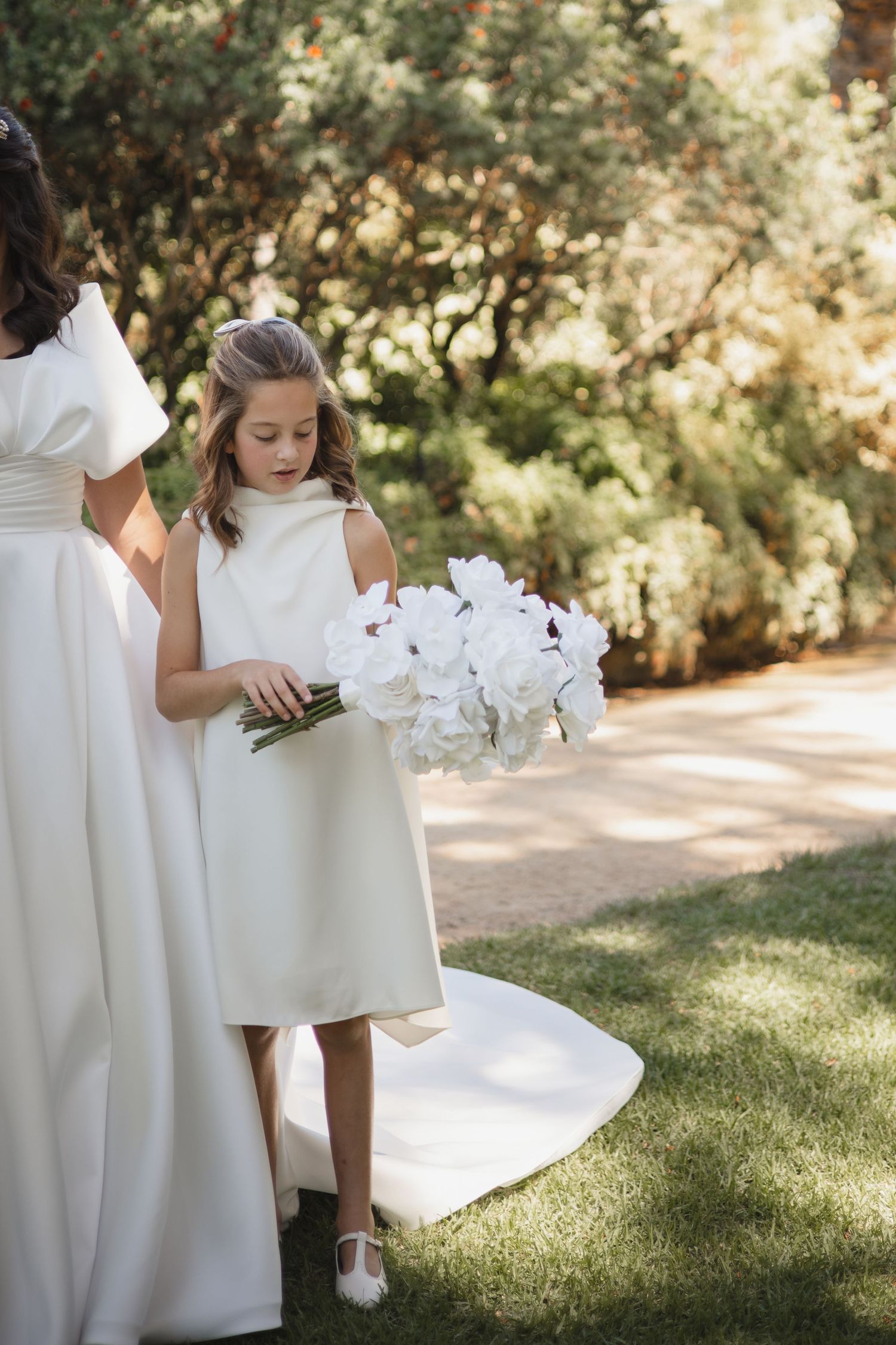 White floral details and elegant dresses create a soft, romantic outdoor wedding scene in dappled sunlight.