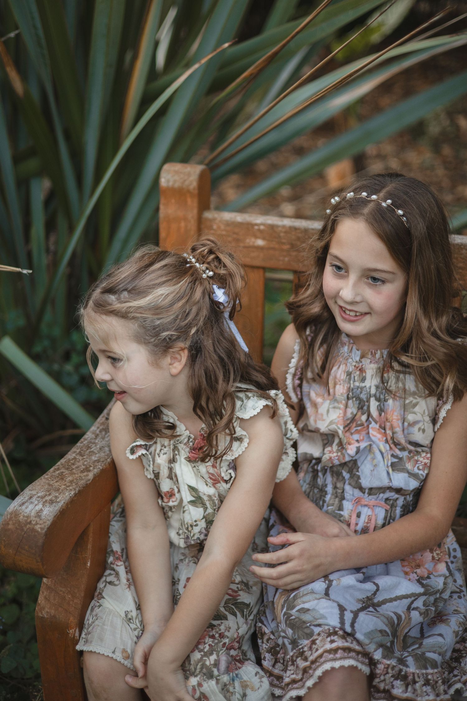 Two children wearing floral dresses sit together on a wooden bench in a garden setting with ornate hair accessories.