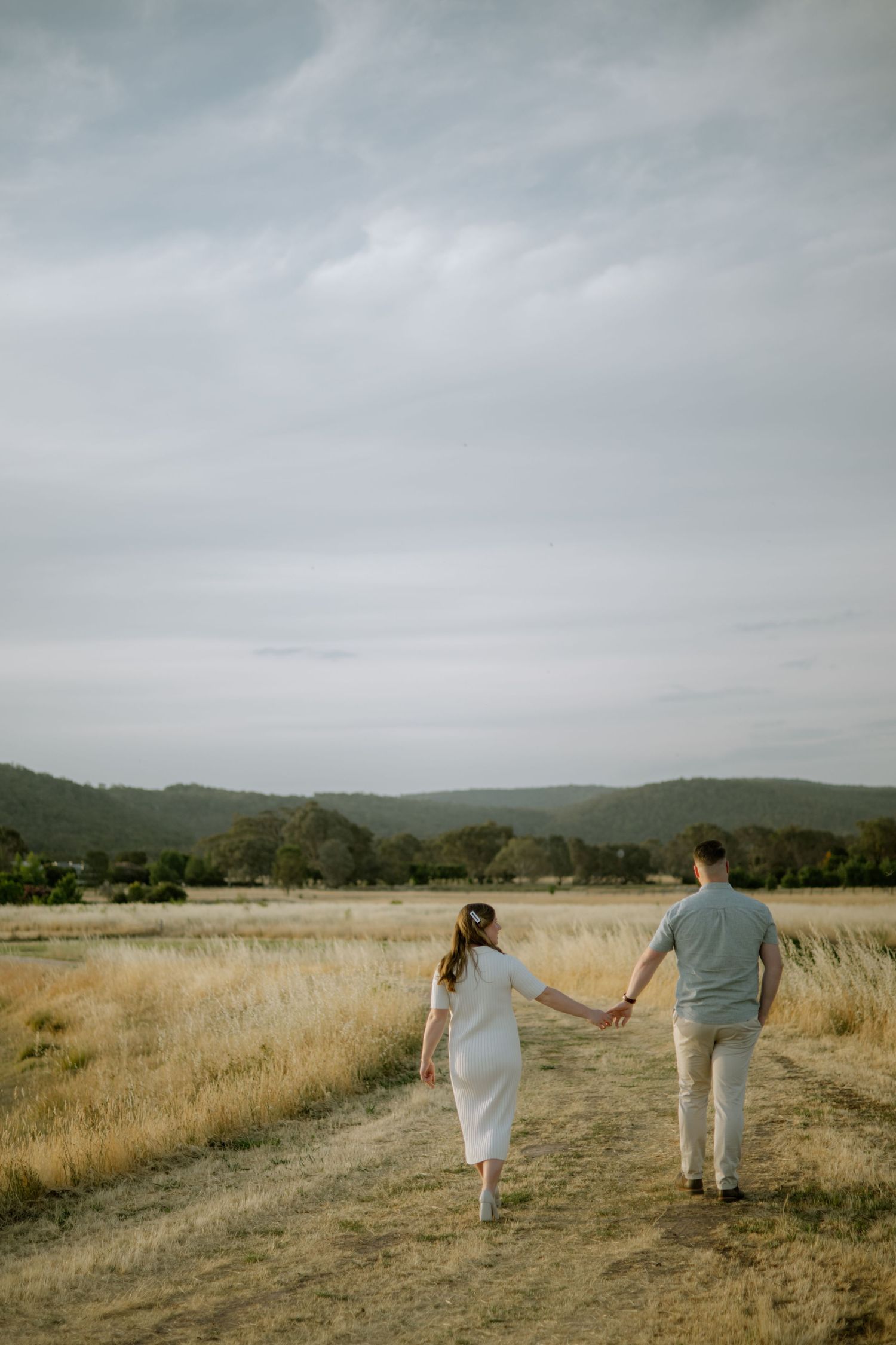 A couple walks hand in hand through a golden grass field on an overcast day with rolling hills in the background.